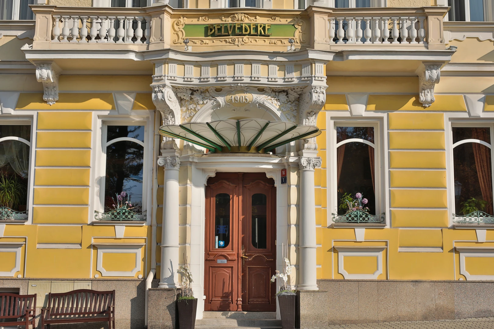 Das Bild zeigt die eindrucksvolle Fassade des Hotel Belvedere Marienbad, die durch ihre historische Architektur und stilvollen Details besticht. Im Zentrum des Bildes befindet sich der Eingangsbereich, der von einem reich verzierten Baldachin in Grün und Weiß geschmückt wird, welcher dem Eingang Eleganz und Schutz verleiht. Über dem Baldachin ist das Baujahr „1905“ prominent in das Steinmauerwerk eingearbeitet, was die historische Bedeutung des Gebäudes unterstreicht. Die gelb-weiße Fassade des Gebäudes ist mit dekorativen Stuckelementen, Säulen und kunstvollen Verzierungen gestaltet, die den klassisch-europäischen Baustil des Hotels hervorheben. Die großen, geschwungenen Fenster sind mit weißen Rahmen und dezenten Eisengittern ausgestattet und geben einen Einblick in die geschmackvoll eingerichteten Innenräume. Vor dem Eingang befinden sich zwei bepflanzte Blumenkübel mit eleganten Orchideen, die den Eingangsbereich einladend gestalten. Neben der Tür laden Holzbanke die Gäste zum Verweilen ein. Der Schriftzug „Belvedere“ in goldenen Lettern auf einem grünen Schild über dem Eingang verleiht der Fassade einen prägnanten und luxuriösen Akzent. Diese prächtige Fassade verkörpert den historischen Charme und den gehobenen Standard, der das Belvedere Marienbad als ein erstklassiges Hotel auszeichnet.