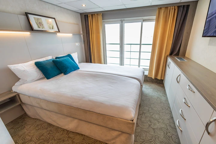 Doppelbett mit weißen Bettlaken und zwei blauen Kissen in einer Kabine mit Balkon, Vorhängen und Kommode mit Schubladen