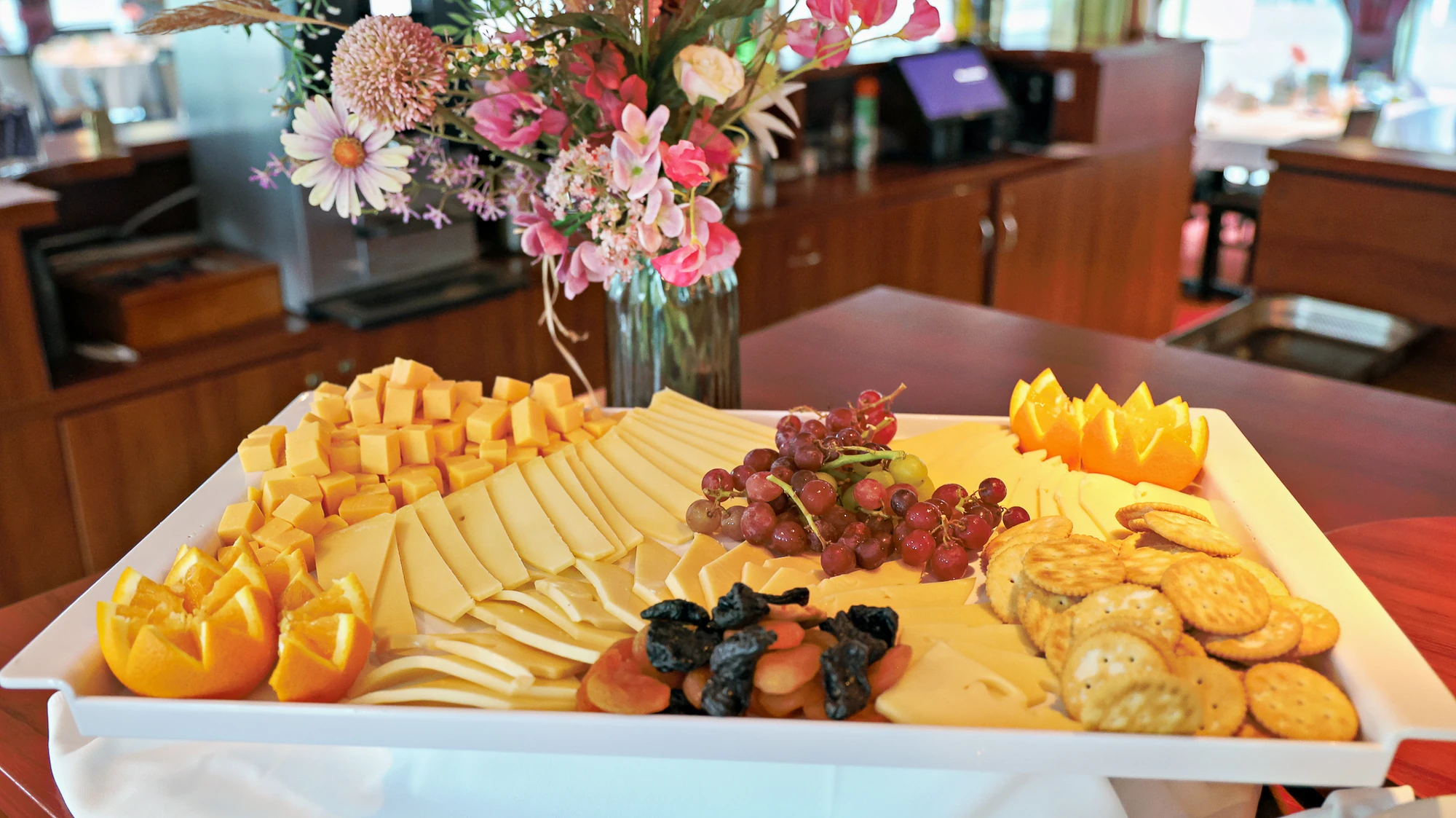 Großes weißes Tablett mit verschiedenen Käsesorten, Trauben, Oliven, Crackern und dekorativen Orangenschnitzen, im Hintergrund Blumenstrauß.