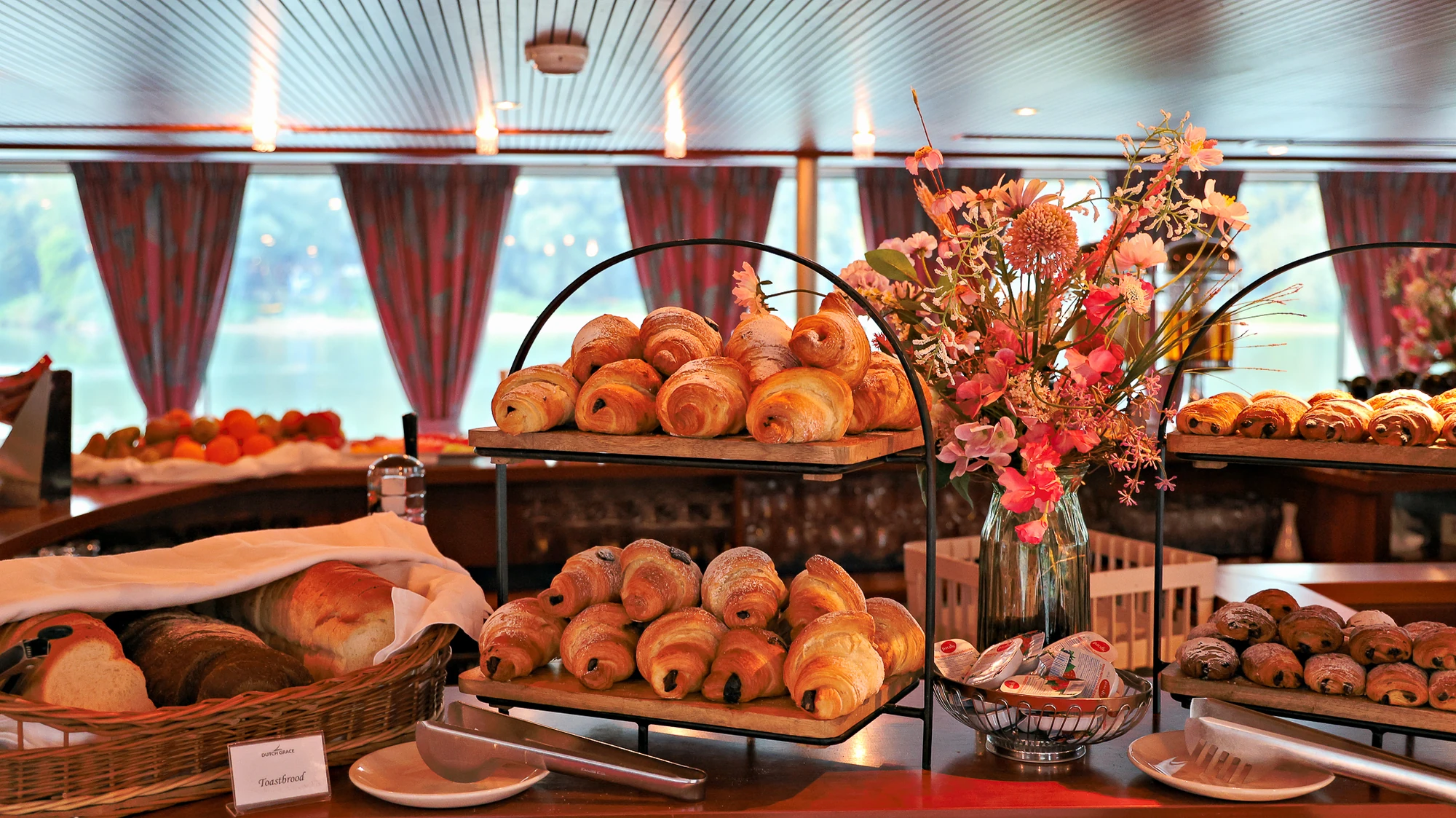 Zweistöckiges Metallgestell mit Croissants und Schokocroissants, daneben Körbe mit Brot und ein Blumenstrauß in einer Glasvase.