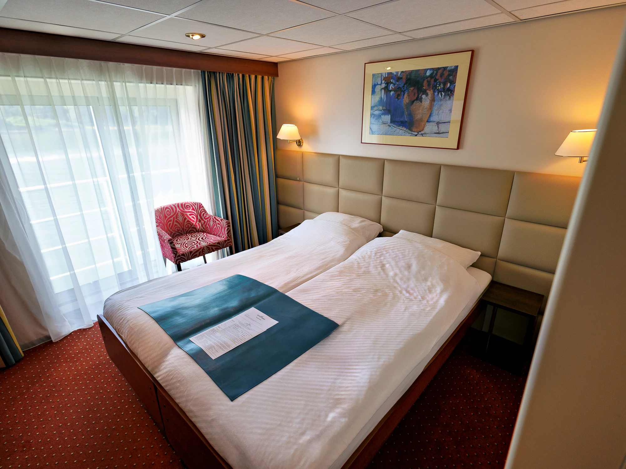 Doppelbett mit weißer Bettwäsche und blauem Läufer, roter Sessel neben bodentiefem Fenster mit Vorhängen, Wandbild und zwei Wandlampen.