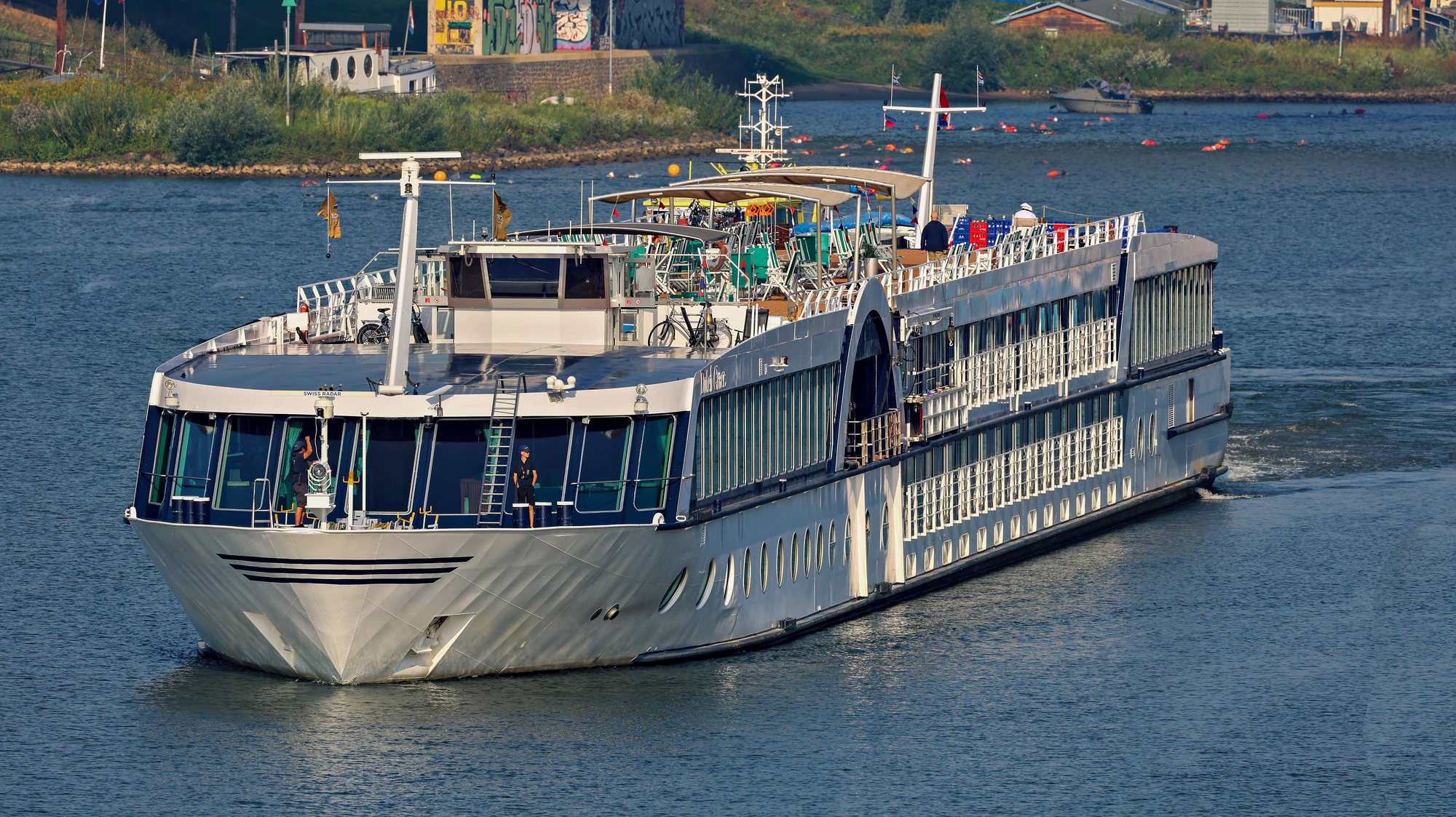 Großes weißes Passagierschiff mit mehreren Decks fährt auf einem Fluss, Ufer mit Gebäuden im Hintergrund.