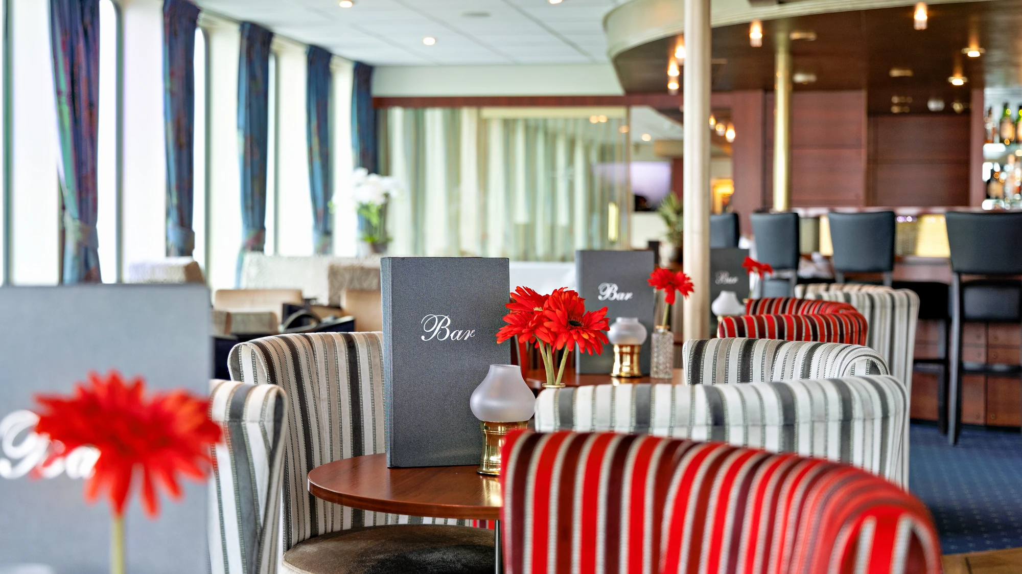Mehrere Tische mit Stühlen in einem hellen Raum, auf einem Tisch steht ein Menü mit der Aufschrift 'Bar' und rote Blumen in einer Vase.