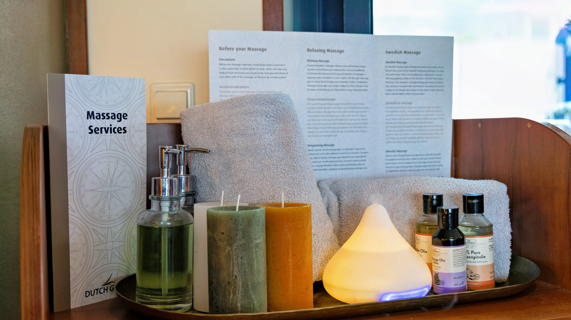 Tablett mit zwei grauen Handtüchern, zwei Kerzen, einem Aromadiffusor, zwei Flaschen Massageöl und einem aufgestellten Flyer mit der Überschrift 'Massage Services'.