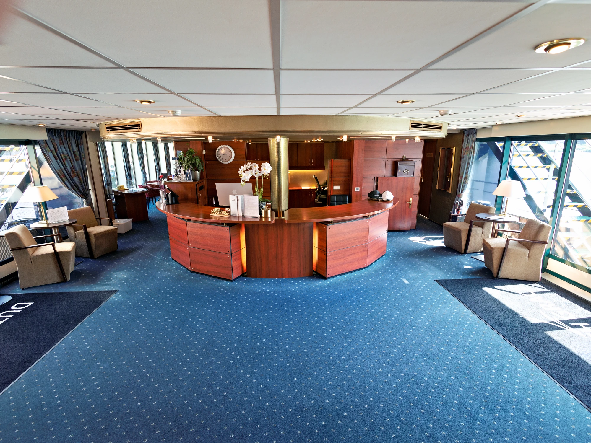 Rezeption mit halbkreisförmigem Holzschalter, blauer Teppichboden, zwei Sesseln und großen Fenstern mit blauen Vorhängen.