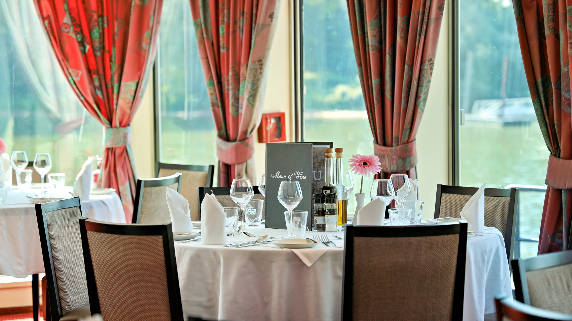 Rund gedeckter Tisch mit weißen Servietten, Weingläsern, Menükarte und rosa Blume vor großen Fenstern mit roten Vorhängen.