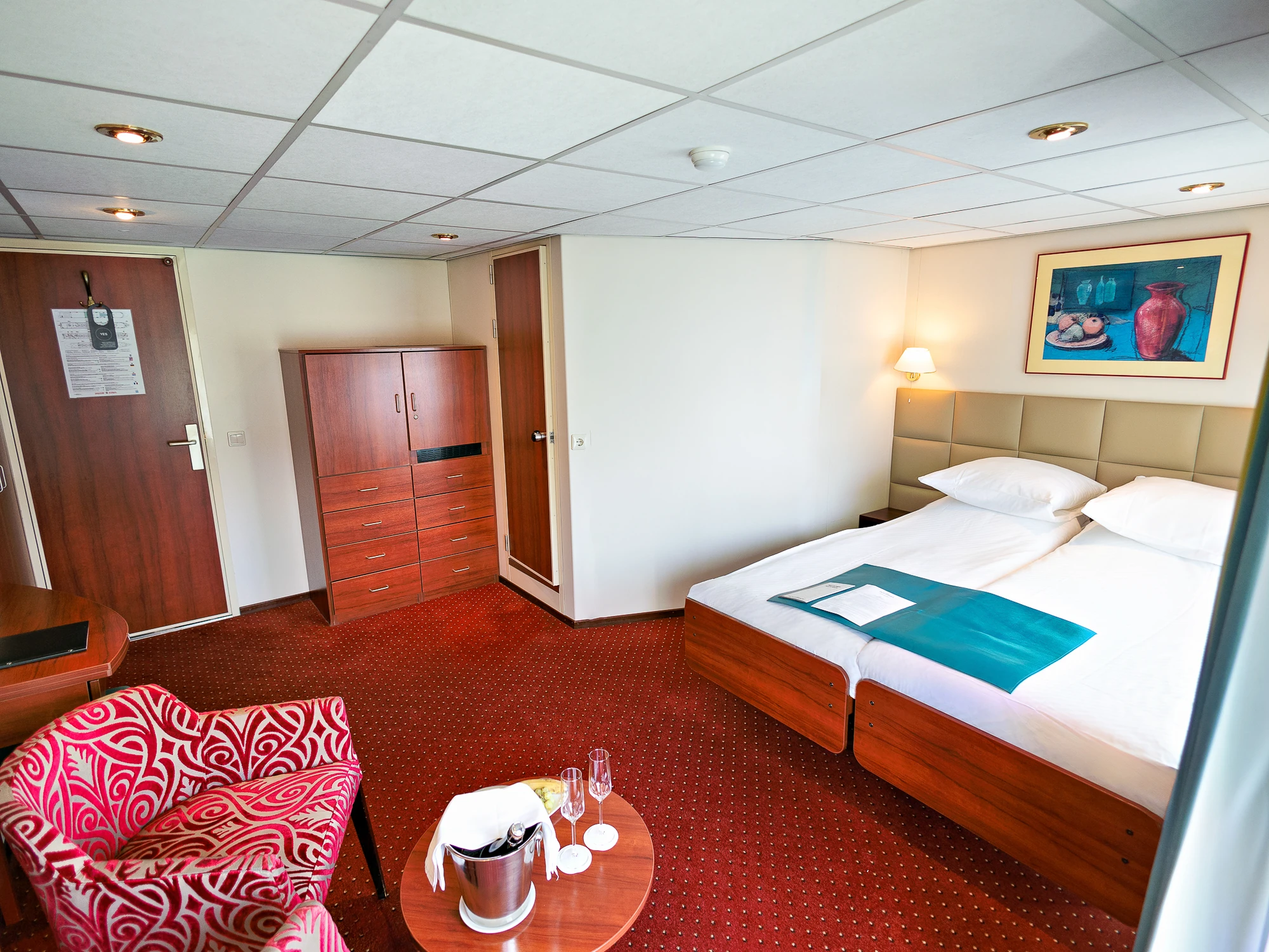Hotelzimmer mit zwei Einzelbetten, rotem Teppich, gemustertem Sessel, kleinem runden Tisch mit Sektglas und Eiskübel, Wandbild und Deckenleuchte.