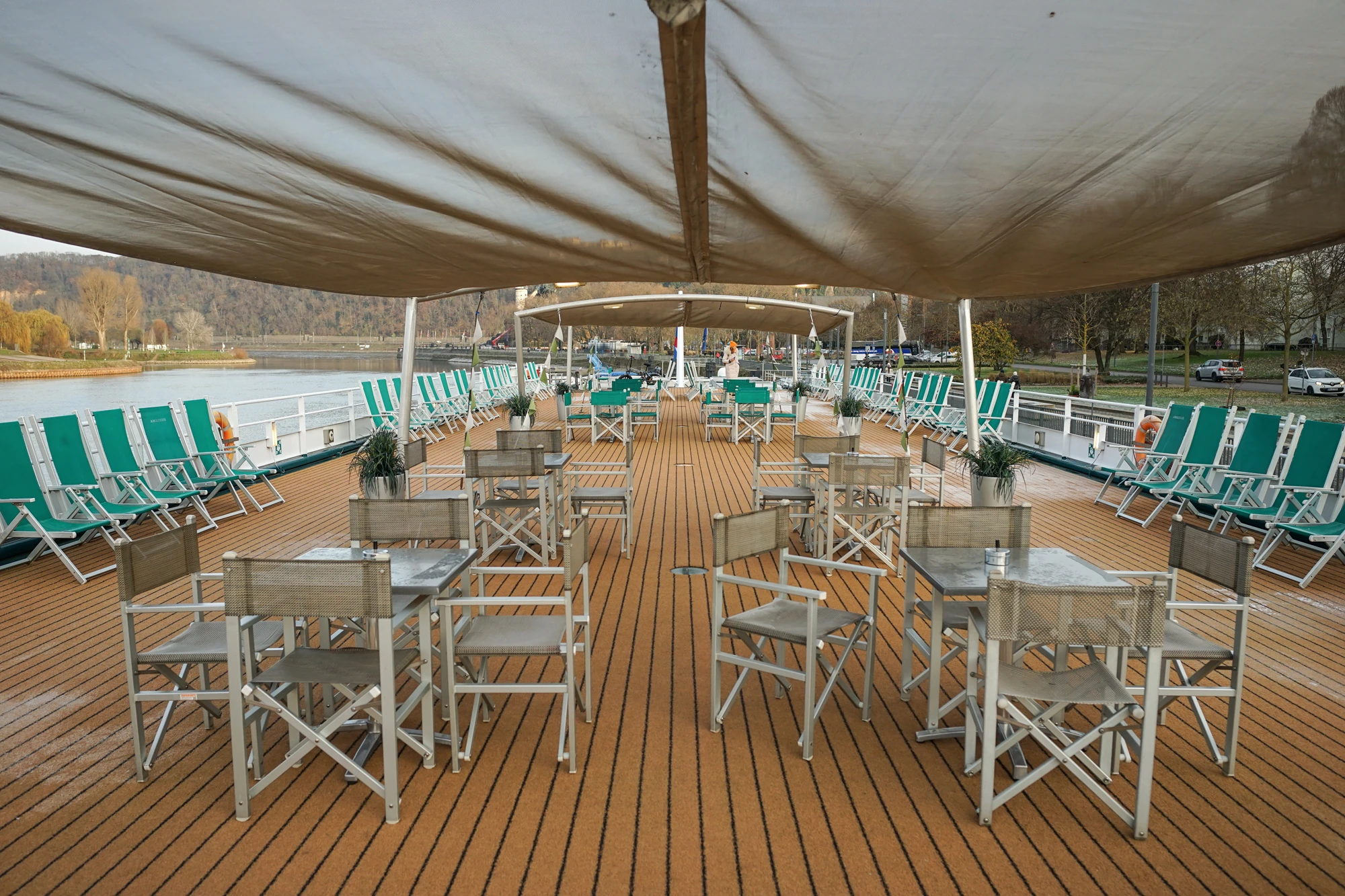 Sonnendach mit mehreren Tischen und Klappstühlen auf dem Holzdeck eines Flusskreuzfahrtschiffs, links und rechts grüne Liegestühle.