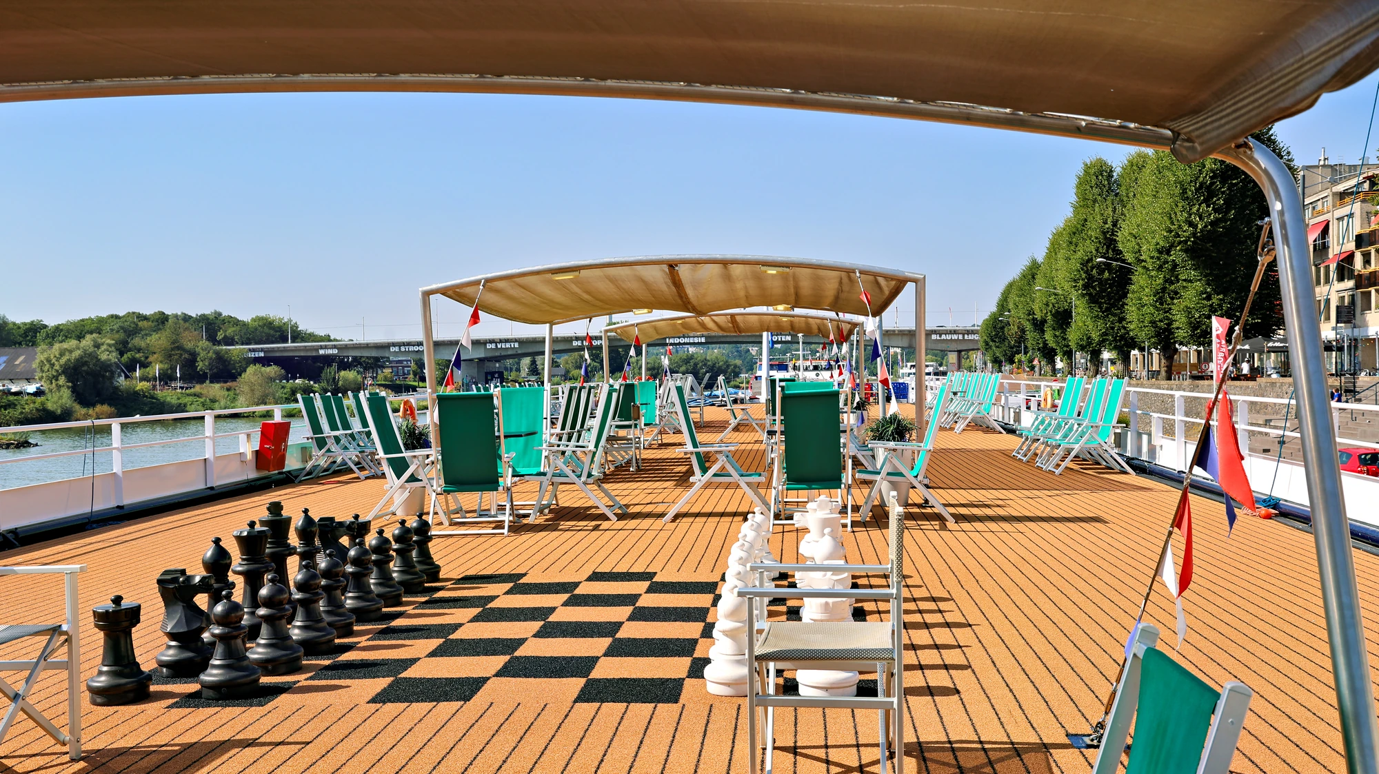 Sonnendeck eines Schiffes mit mehreren grünen Liegestühlen, einem großen Schachspiel mit schwarzen und weißen Figuren und einem Sonnensegel.