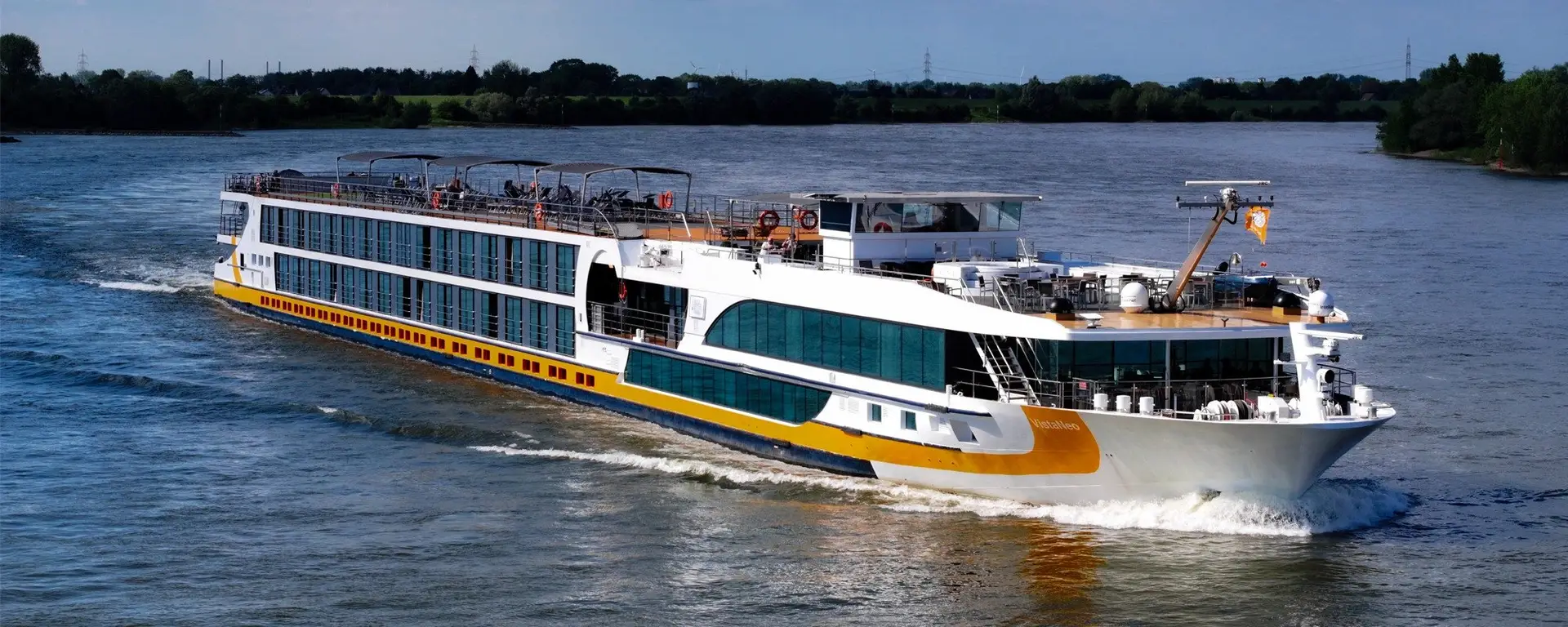 Flusskreuzfahrtschiff mit mehreren Decks und großen Fenstern fährt auf einem breiten Fluss unter bewölktem Himmel.