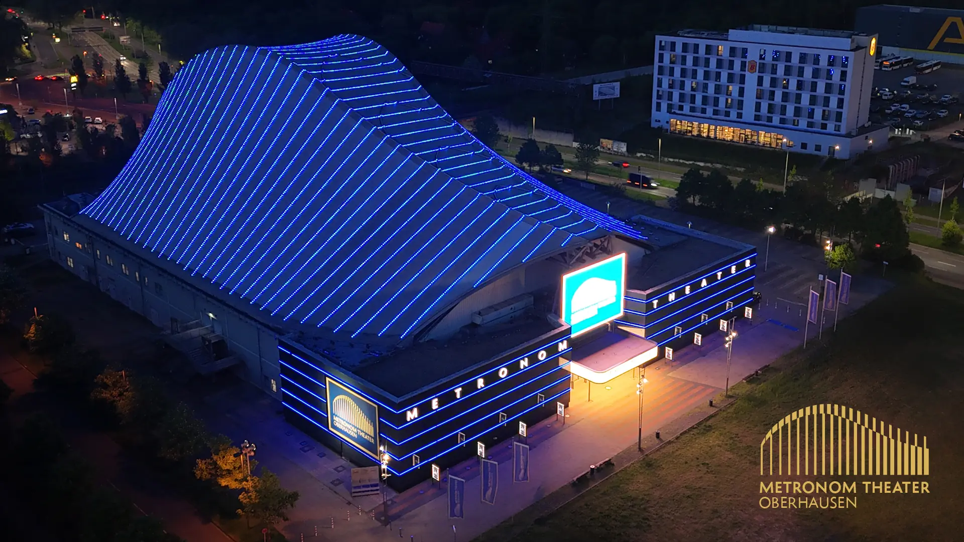 Metronom Theater Oberhausen von außen: Eingang des Metronom Theaters in Oberhausen bei Abenddämmerung in blauer Beleuchtung und Schriftzug 'Metronom Theater' sowie Willkommensbanner