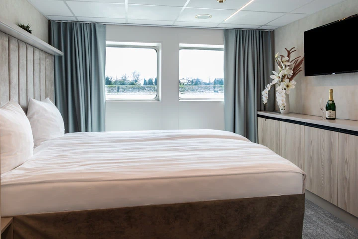 Doppelbett mit weißen Kissen und Bettdecke vor zwei Fenstern mit grauen Vorhängen in einer Kabine, rechts Sideboard mit Fernseher, Champagnerflasche und Blumendekoration