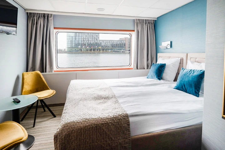 Kabine mit zwei Einzelbetten, blauen Kissen, grauer Wand, gelbem Stuhl und Fenster mit grauen Vorhängen