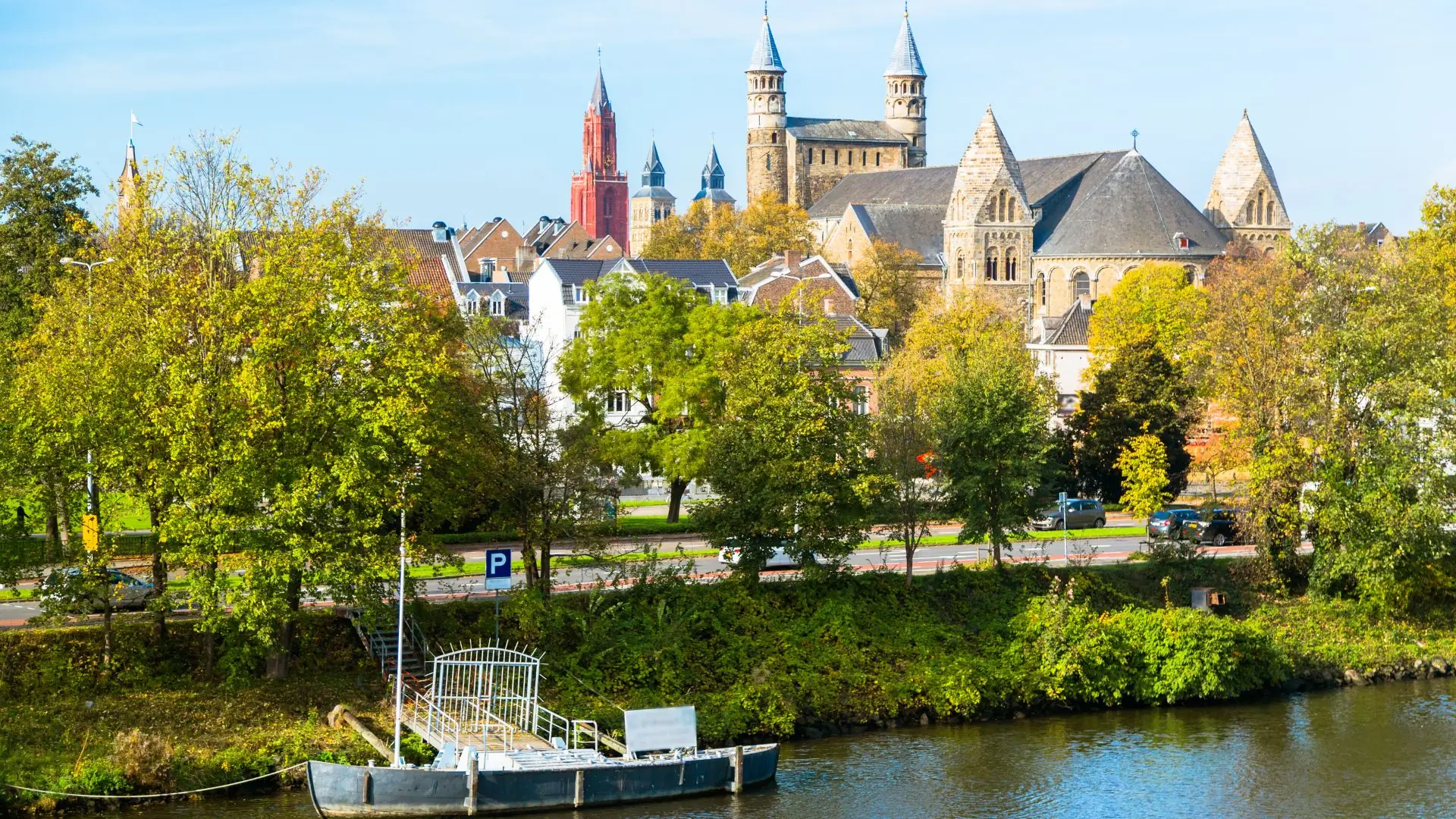 Blick auf Maastricht mit der Basilika des Heiligen Servatius und der Sint-Janskerk mit rotem Turm, Bäume und Wasser im Vordergrund.