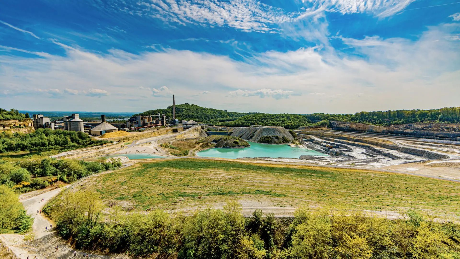Landschaft mit grünem Gras, Büschen und Teichen vor Industrieanlagen der ENCI-Steingrube Maastricht unter blauem Himmel mit Wolken.