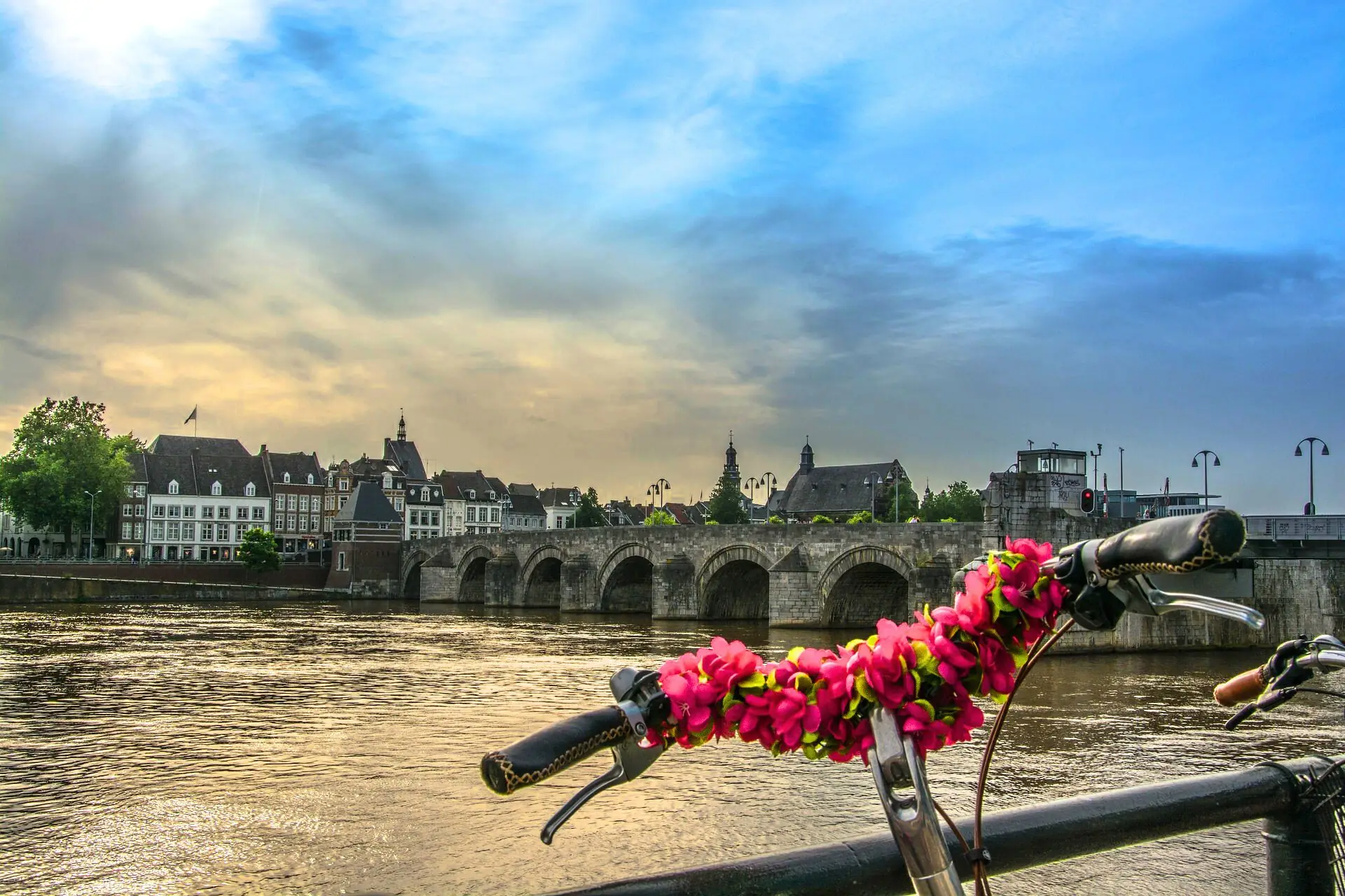 Fahrradlenker mit pinker Blumenkette vor der historischen Steinbrücke und Gebäuden in Maastricht bei bewölktem Himmel.
