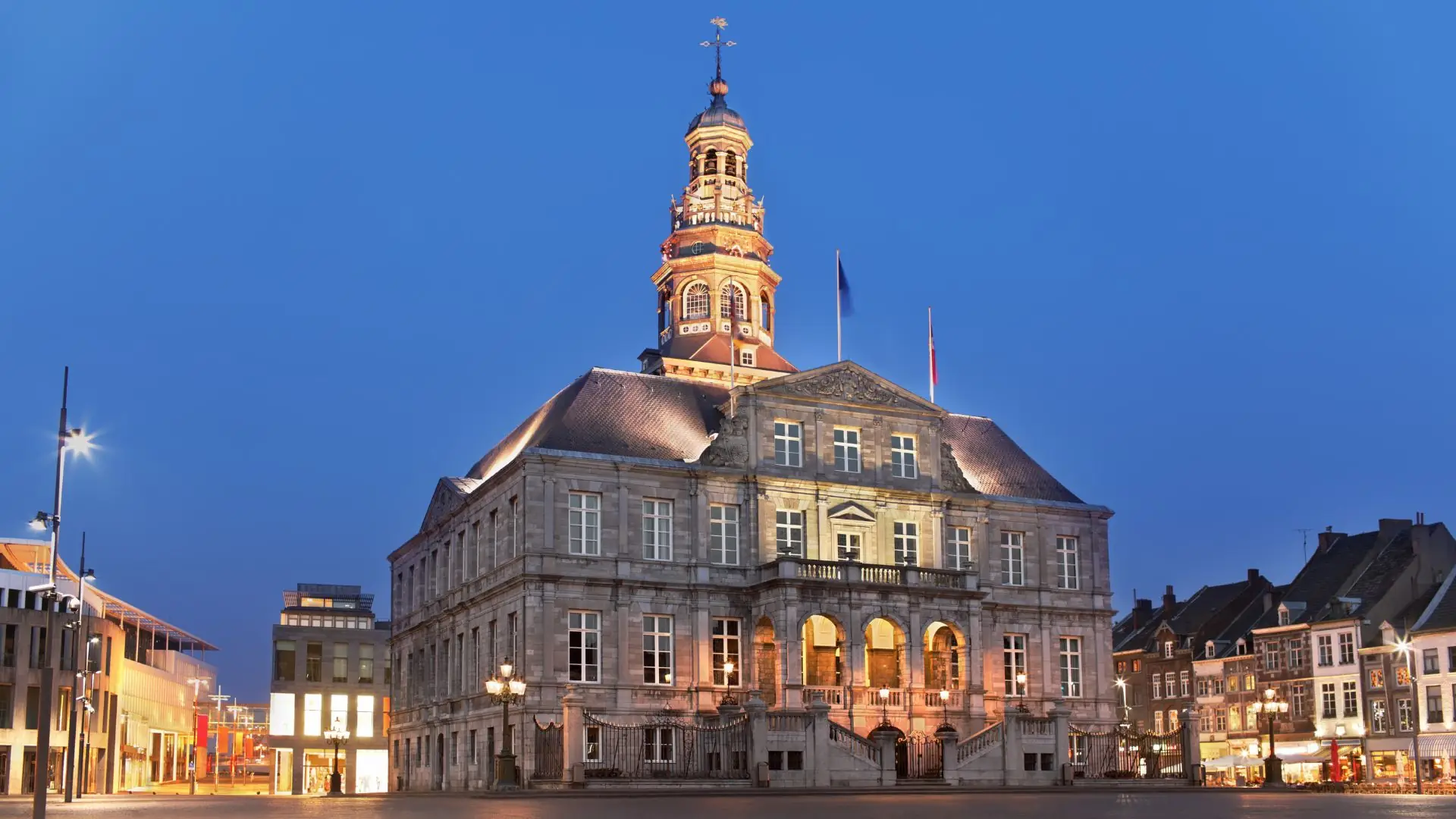 Beleuchtetes historisches Rathaus von Maastricht mit Turm und mehreren Fenstern bei Abenddämmerung.