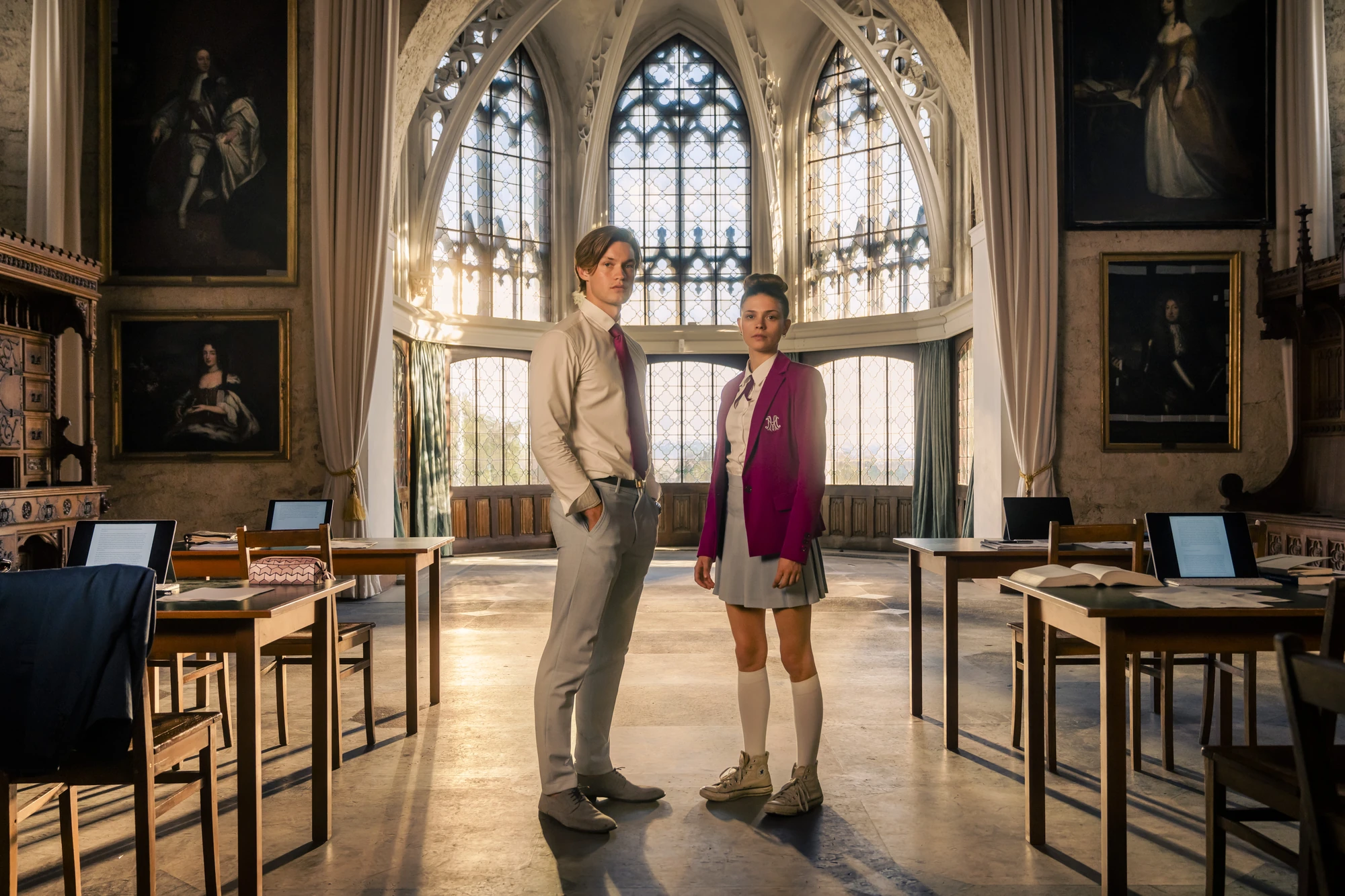 Maxton Hall Hannover Schloss Marienburg zeigt Ruby und James in einem eleganten, historischen Klassenzimmer mit gotischen Fenstern, umgeben von antiken Möbeln und Kunstwerken.