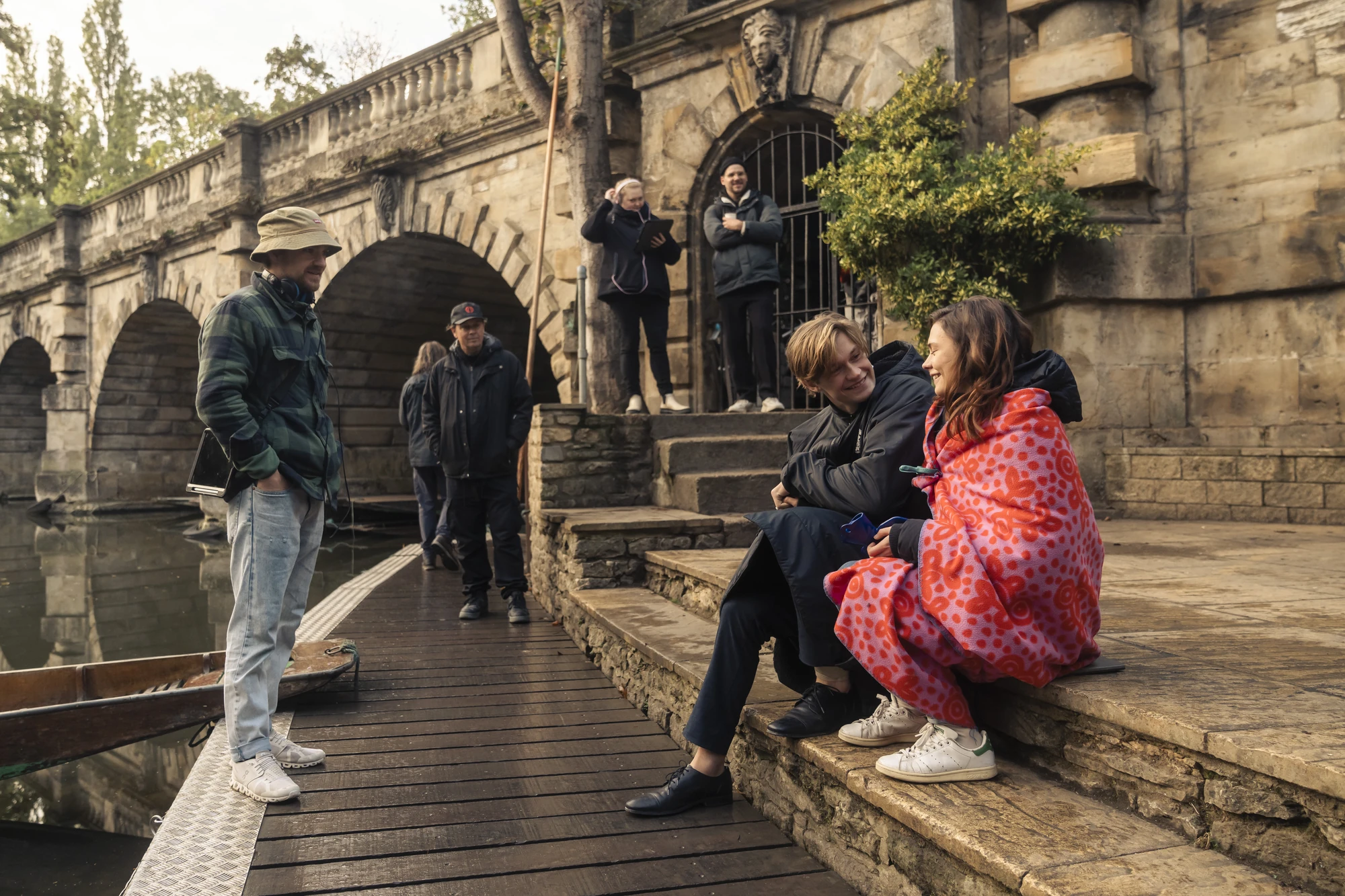 Maxton Hall Dreharbeiten zeigt Ruby und James, eingehüllt in eine warme Decke, die auf den Steinstufen am Wasser sitzen, umgeben von der Filmcrew vor der Kulisse eines historischen Gebäudes.