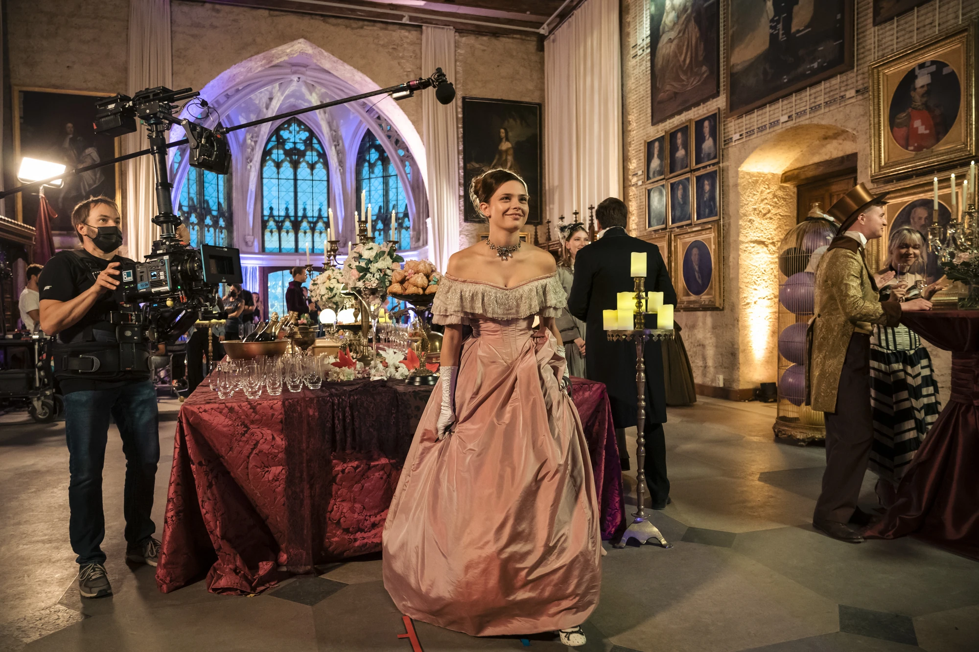 Eine Szene aus Maxton Hall mit Lydia in einem eleganten Ballkleid. Hinter ihr die aufwendig dekorierte Kulisse und Crewmitglieder mit Kamera- und Tonausrüstung.
