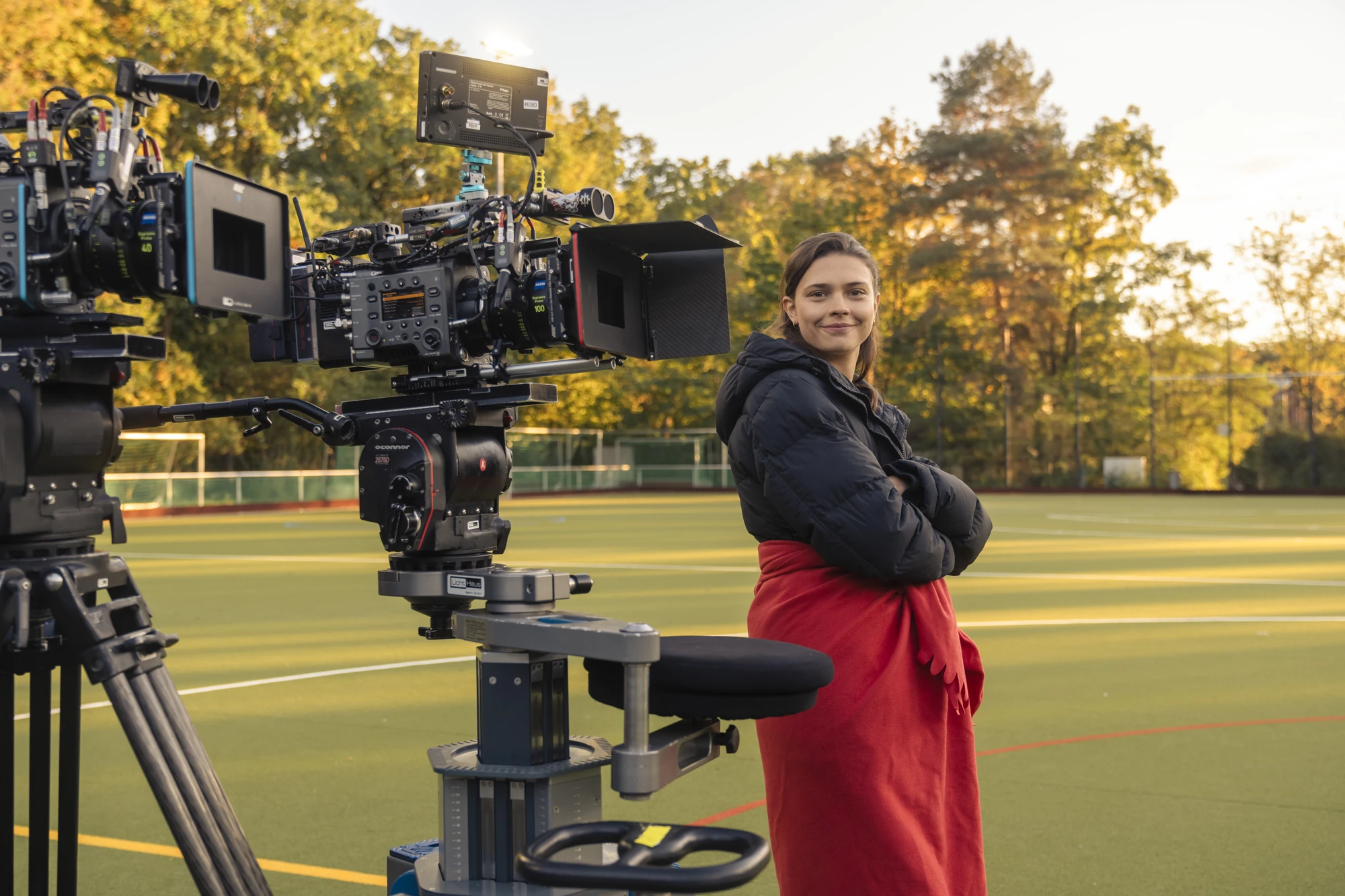 Maxton Hall Dreharbeiten zeigt Ruby in warmer Kleidung, lächelnd vor einer Filmkamera auf einem Sportplatz, umgeben von herbstlichen Bäumen und unter einem klaren Himmel.