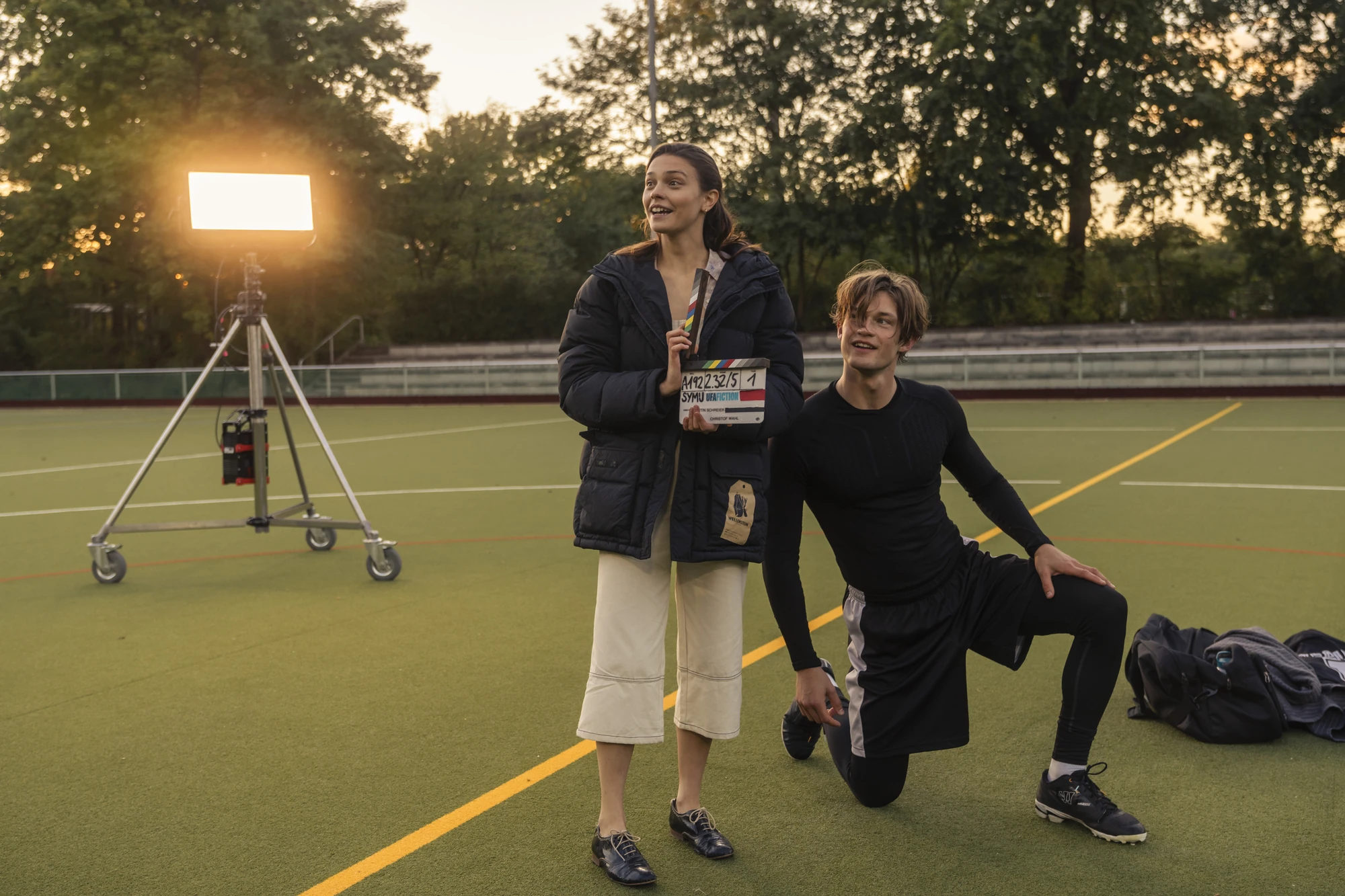 Maxton Hall Dreharbeiten zeigt Ruby mit einer Filmklappe in der Hand und James in Sportkleidung auf einem Sportplatz, beide lächelnd in einer fröhlichen Filmszene.