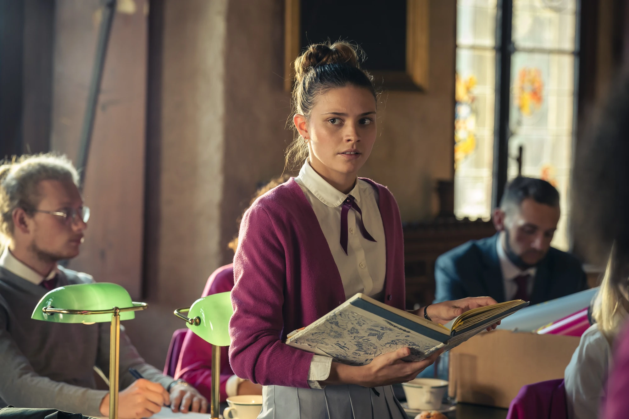 Maxton Hall Szenenbild zeigt Ruby im historischen Klassenzimmer offensichtlich überrascht oder verwundert, mit einem alten Buch in der Hand. Die Szene vermittelt eine Atmosphäre von Spannung und Geheimnissen.