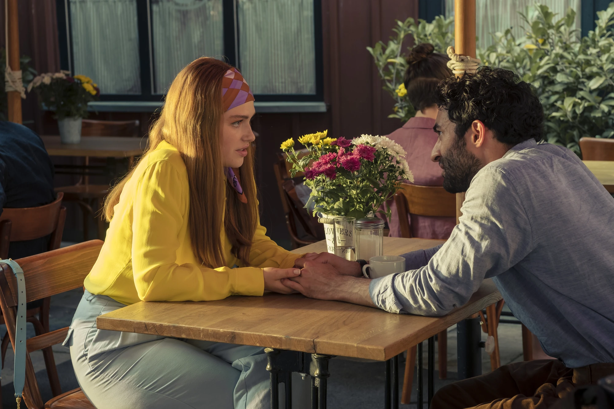 Maxton Hall Szenenbild zeigt eine intensive Szene zwischen Lydia und dem Lehrer , die sich an einem Tisch in einem gemütlichen Café gegenüber sitzen, Hände haltend und in ein tiefes Gespräch vertieft sind.