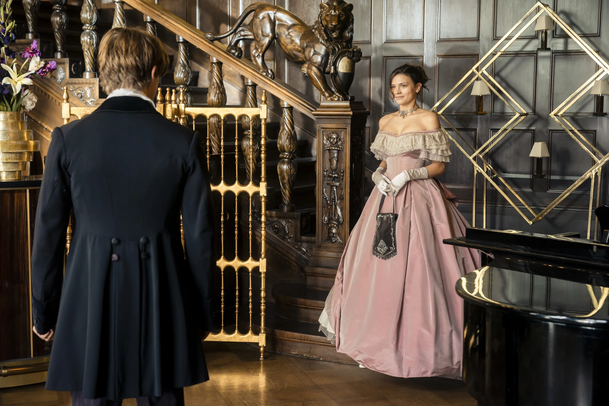 Maxton Hall Szenenbild - Grace in einem eleganten Ballkleid begegnet einen jungen Mann auf einer prunkvollen Treppe im Maxton Hall.