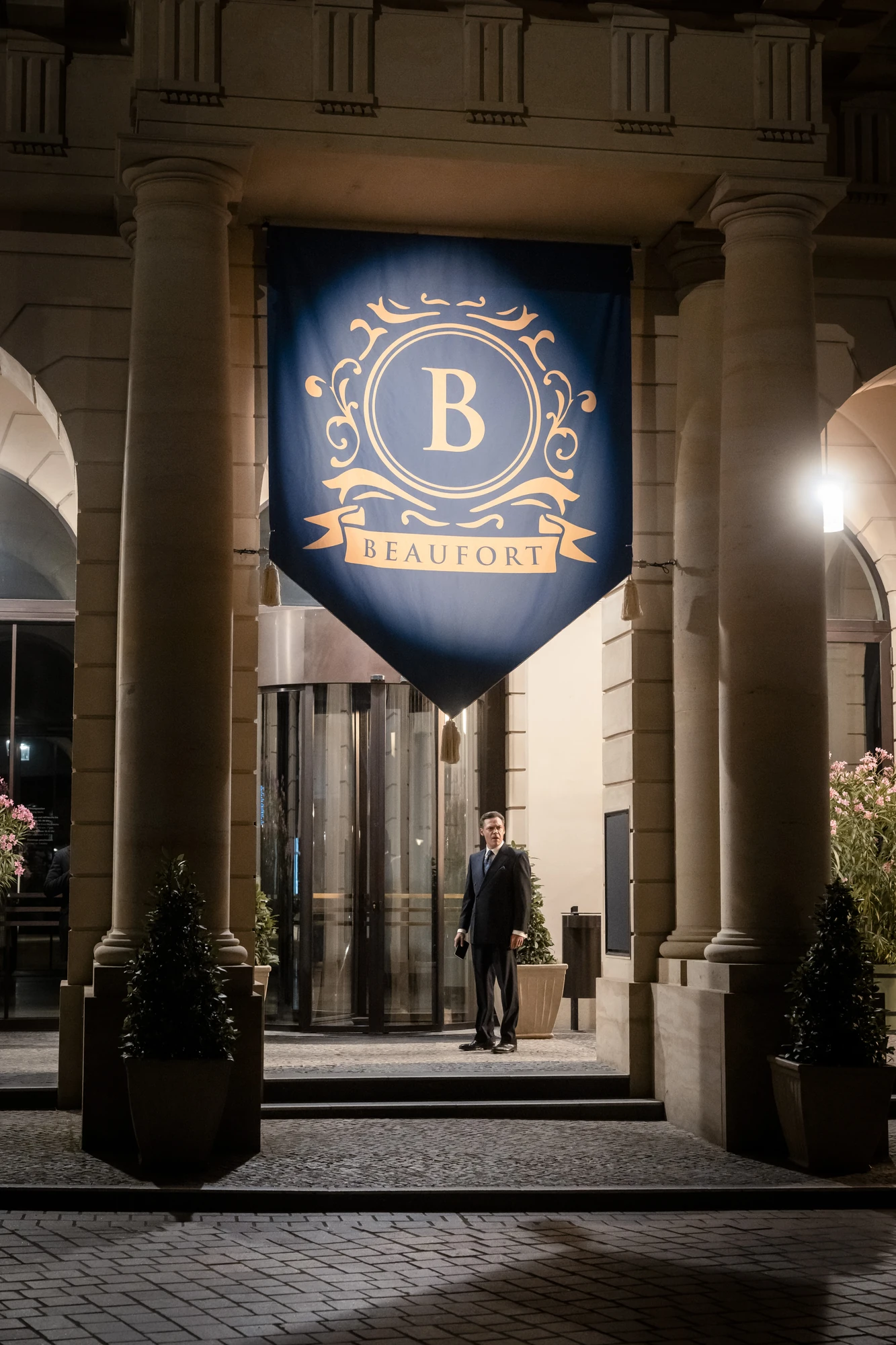 Maxton Hall Szenenbild - ein Mann steht vor dem Beaufort Gebäude. Das Beaufort Wappen in Blau hängt vom Gebäude runter.