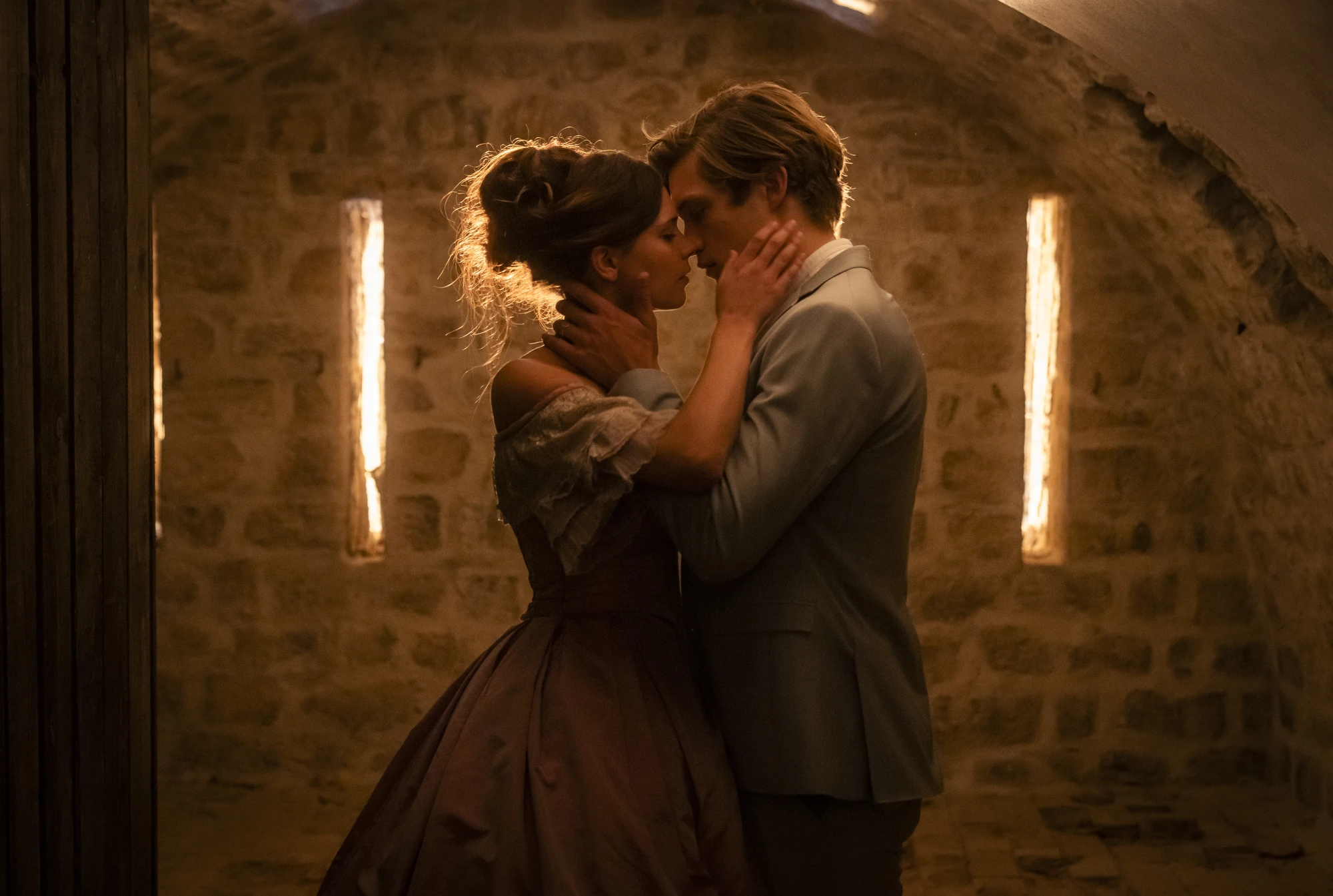 Maxton Hall Szenenbild zeigt einen intimen Moment zwischen Ruby und James, die sich in eleganter Abendgarderobe in einem stimmungsvoll beleuchteten Steingewölbe leidenschaftlich näherkommen.