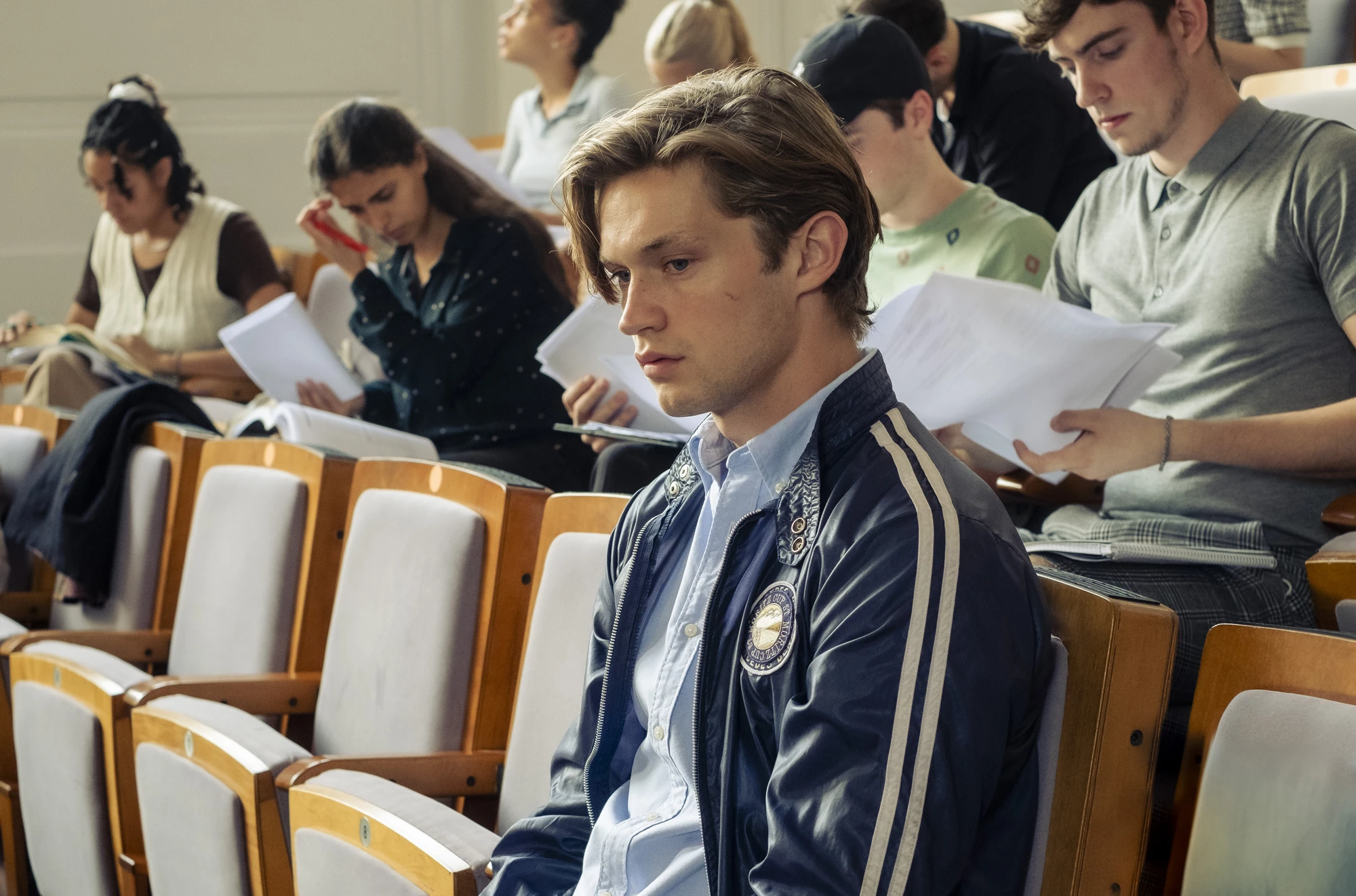 Maxton Hall Szenenbild - James sitzt nachdenklich in einem Hörsaal, umgeben von anderen Schülern. Eine Momentaufnahme, die innere Konflikte und die Intensität des Schulalltags bei Maxton Hall zeigt.