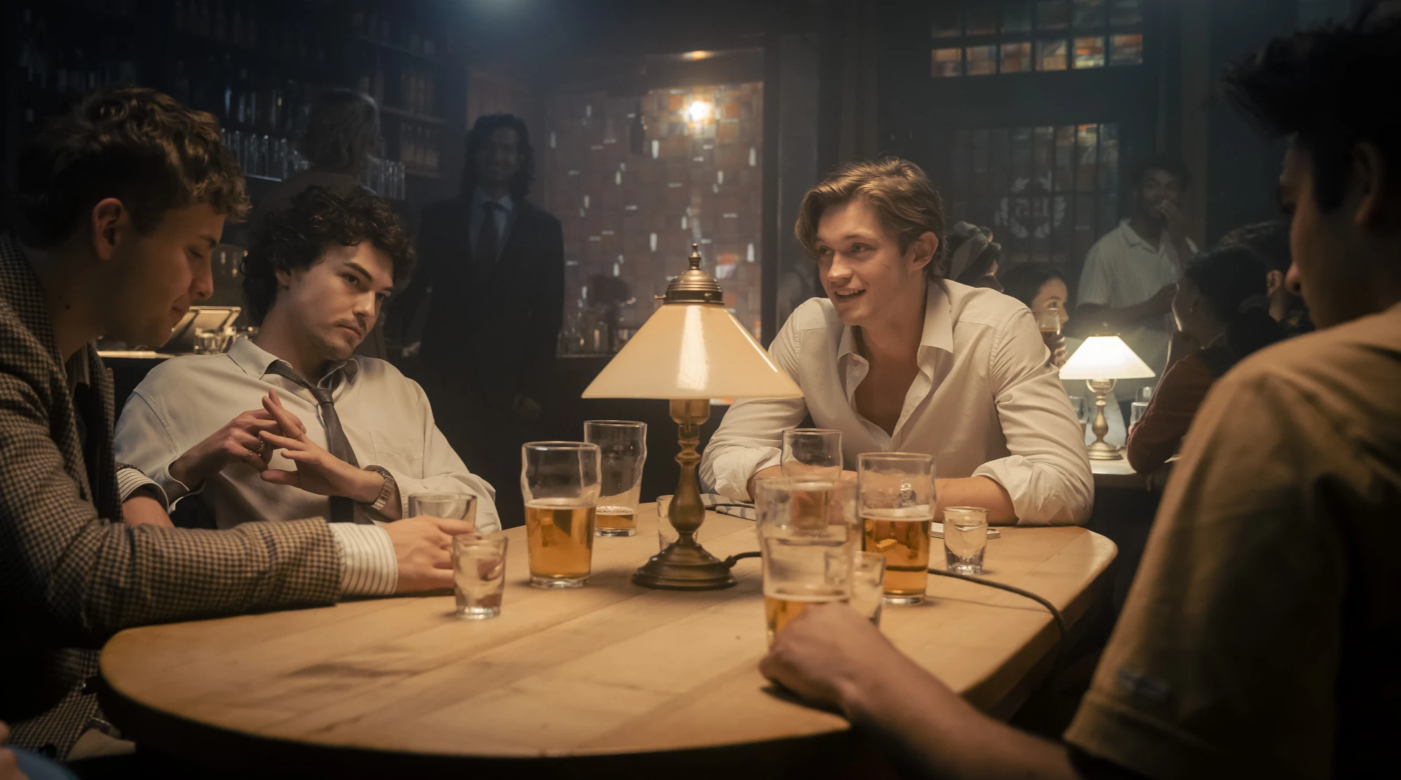 Maxton Hall Szenenbild - James Beaufort und weitere Charaktere am Tisch am reden und trinken dabei.