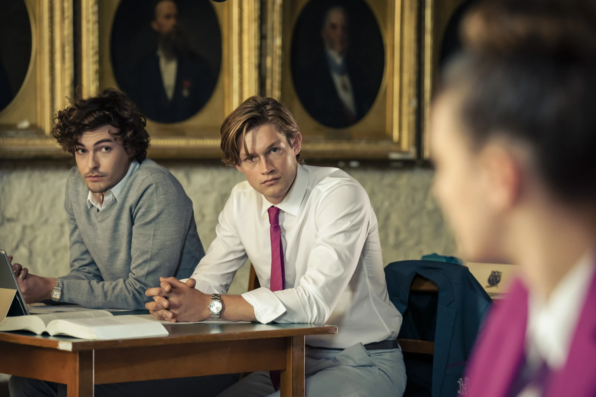 Maxton Hall zeigt James und einen weiteren Schüler in einem klassischen Unterrichtsraum, konzentriert sitzend, umgeben von historischen Porträts an der Wand und James und Ruby haben Blickkontakt.