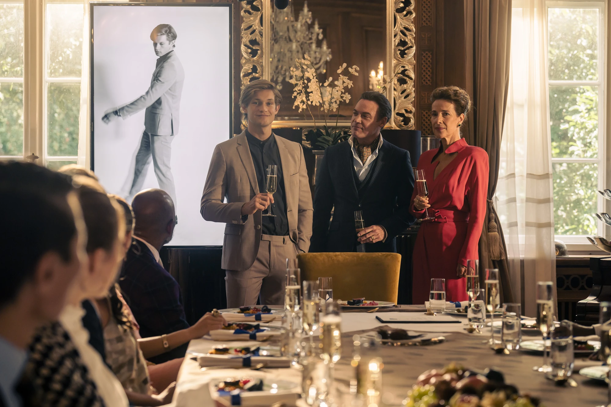 Maxton Hall zeigt James bei einem eleganten Abendessen. Er steht mit einem Glas Sekt in der Hand, flankiert von zwei Erwachsenen in festlicher Kleidung, während die Gäste aufmerksam zusehen.