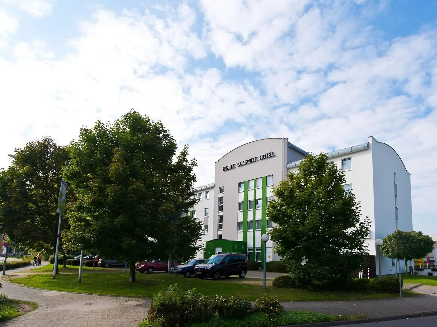 Das ACHAT Hotel Monheim am Rhein mit weißer Fassade, grünem Eingangsbereich und mehreren parkenden Autos davor, umgeben von Bäumen und einem Gehweg im Vordergrund
