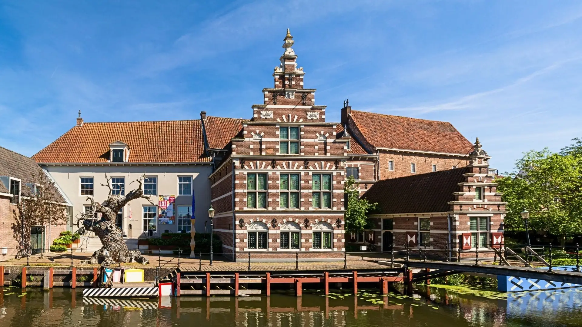 Historisches Backsteingebäude mit Treppengiebel und grünen Fensterläden am Wasser, davor Brücke und Baumskulptur, Museum Flehite in Amersfoort.