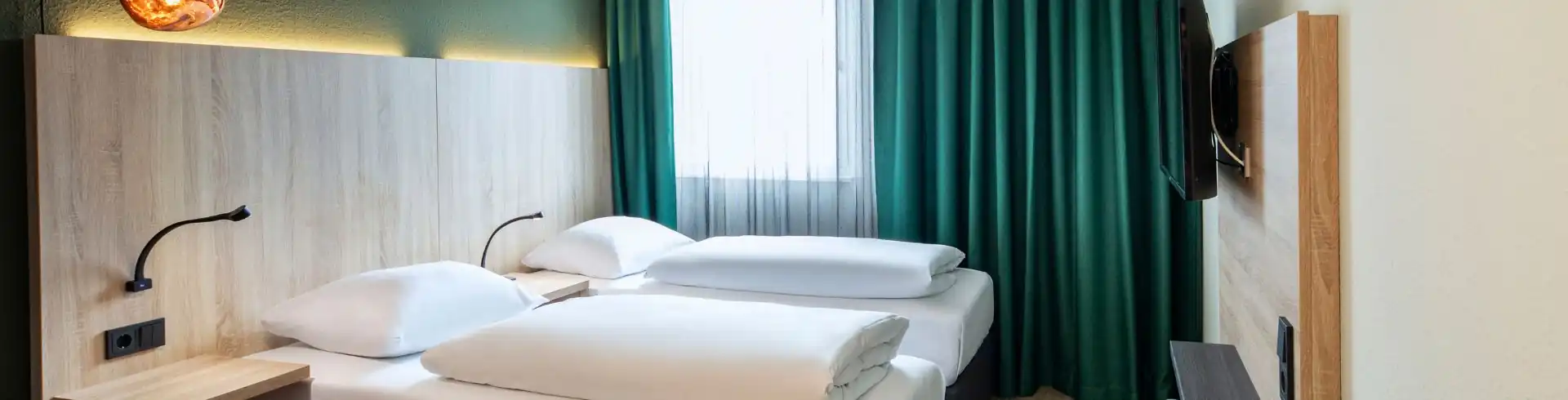 Banner Hotelzimmer mit zwei Einzelbetten, Nachttischlampen, grünem Vorhang, Wandfernseher und zwei roten Stühlen