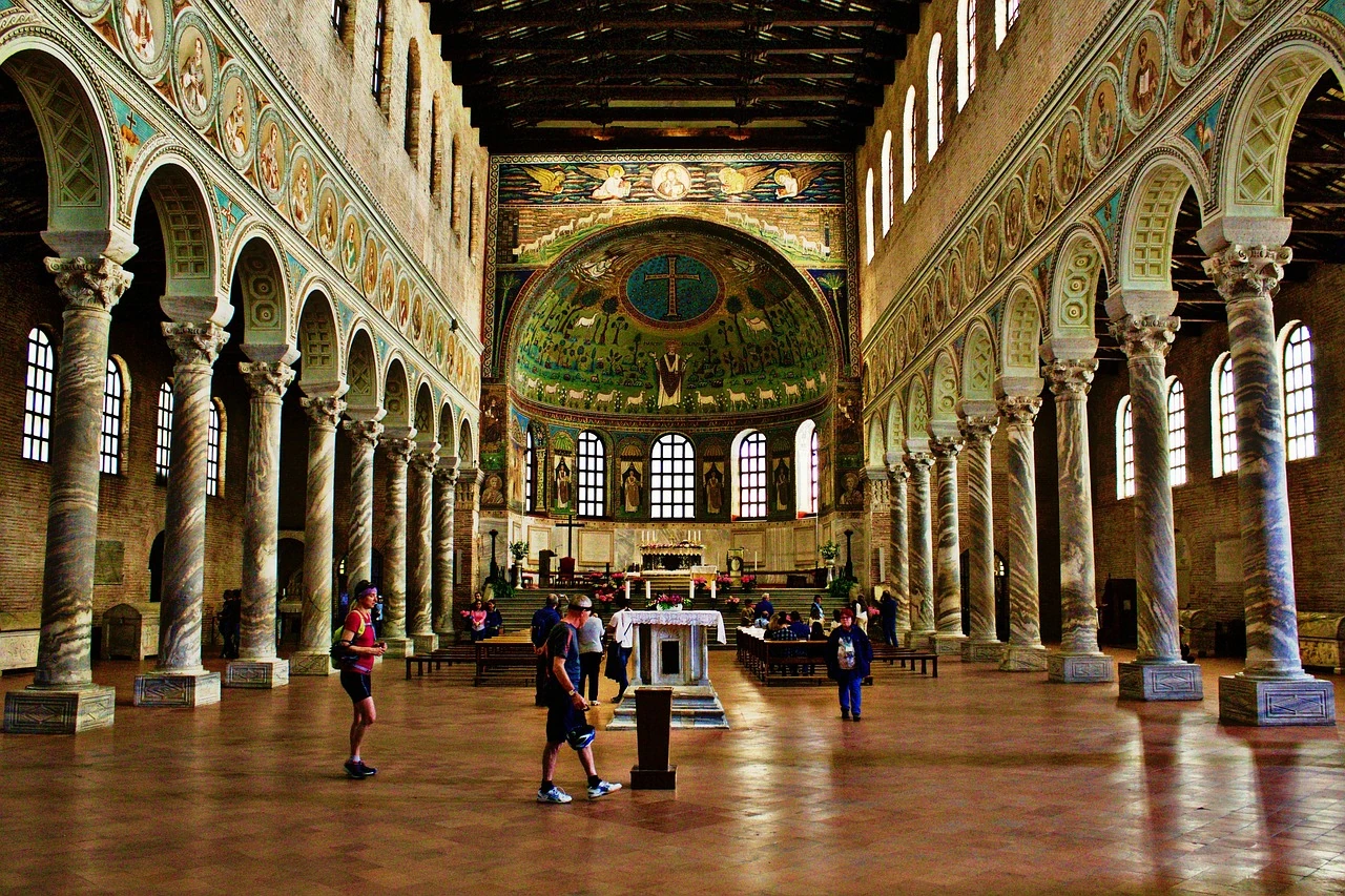 Innenansicht der San Vitale Basilika in Ravenna, die für ihre prächtigen Mosaike und beeindruckende Architektur bekannt ist. Besucher bewundern die farbenfrohen Kunstwerke und die kunstvoll gestalteten Bögen, die dem Raum eine spirituelle und majestätische Atmosphäre verleihen.