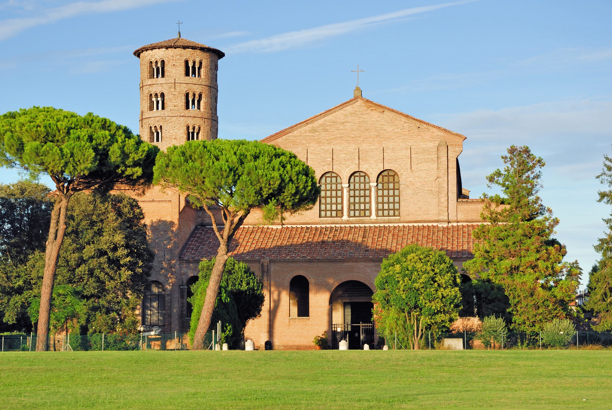 Die Basilika Sant’Apollinare in Classe erstrahlt bei sonnigem Tageslicht, umgeben von einer gepflegten Parklandschaft. Die markante Architektur der Basilika fügt sich harmonisch in die grüne Umgebung ein und schafft eine friedliche, einladende Szenerie.