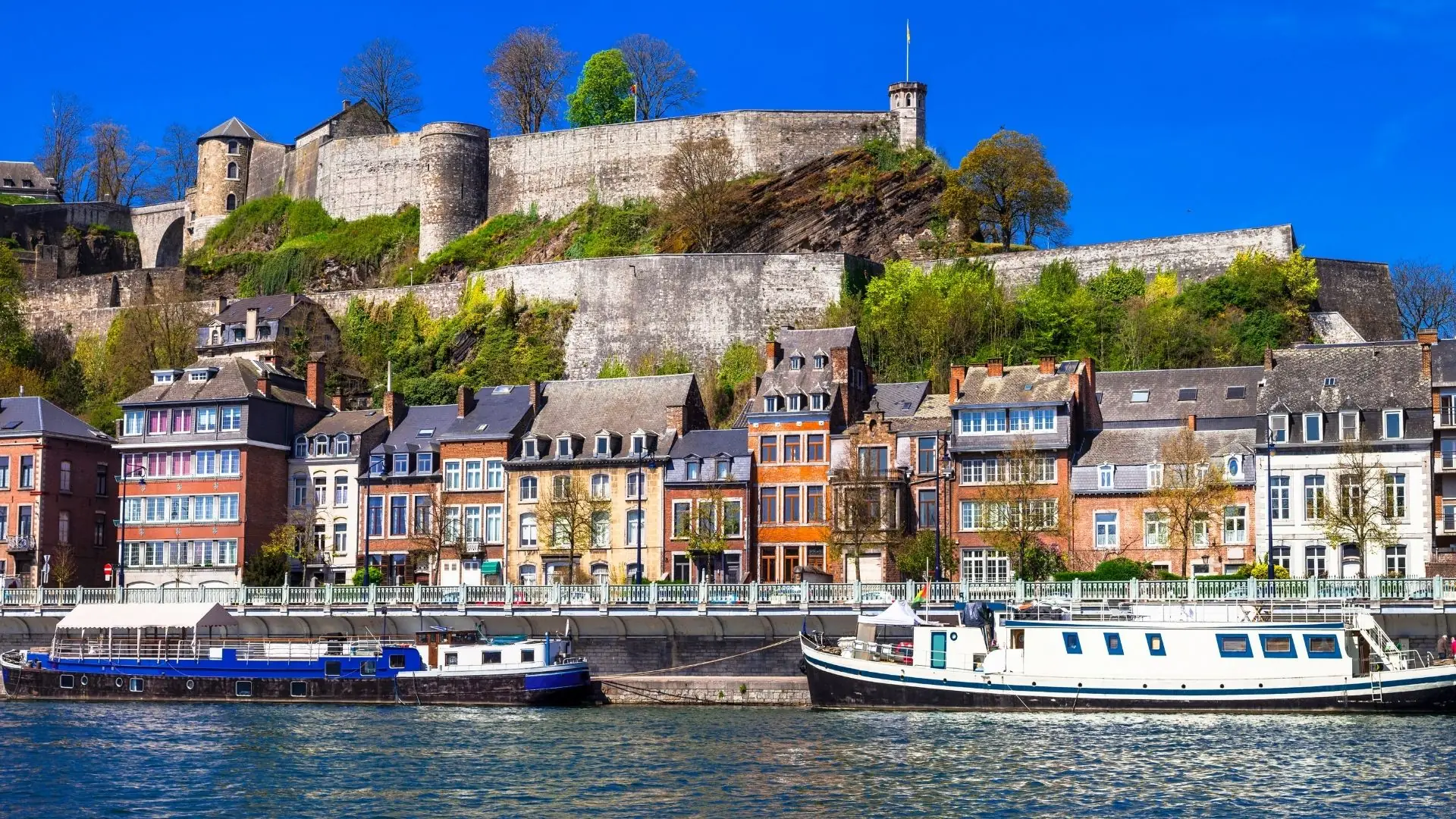 Bunte Häuserzeile am Flussufer mit zwei angelegten Booten und einer Burg auf einem bewaldeten Hügel im Hintergrund