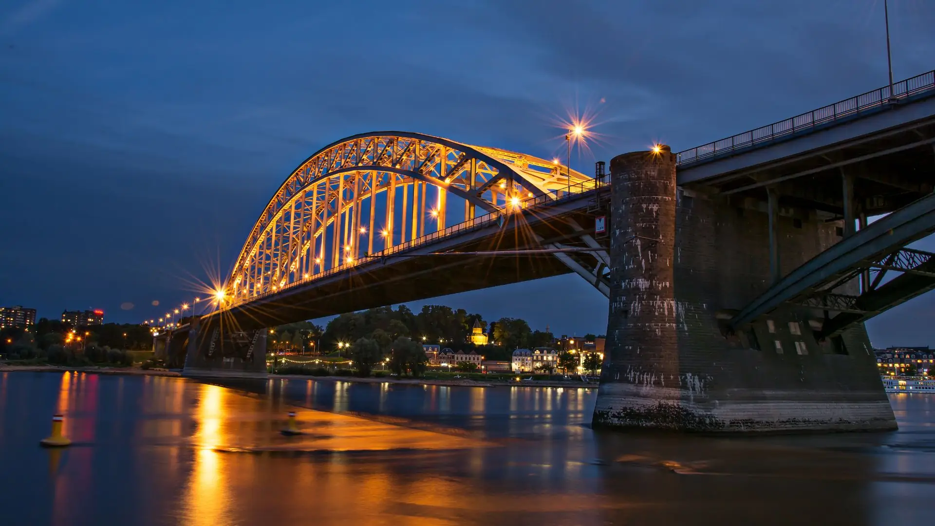 Beleuchtete Eisenbogenbrücke über einen Fluss bei Nacht in Nijmegen, mit reflektierendem Wasser und Stadtlichtern im Hintergrund