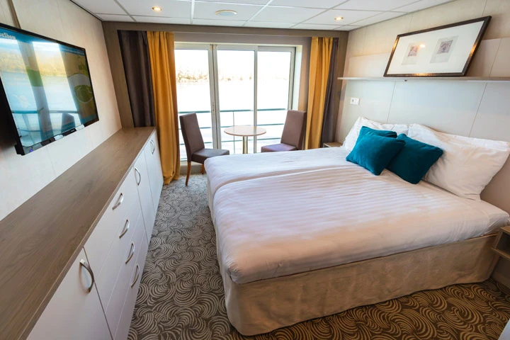 Kabine mit Doppelbett, zwei blauen Kissen, zwei Stühlen und kleinem Tisch vor bodentiefen Fenstern mit Blick auf Wasser