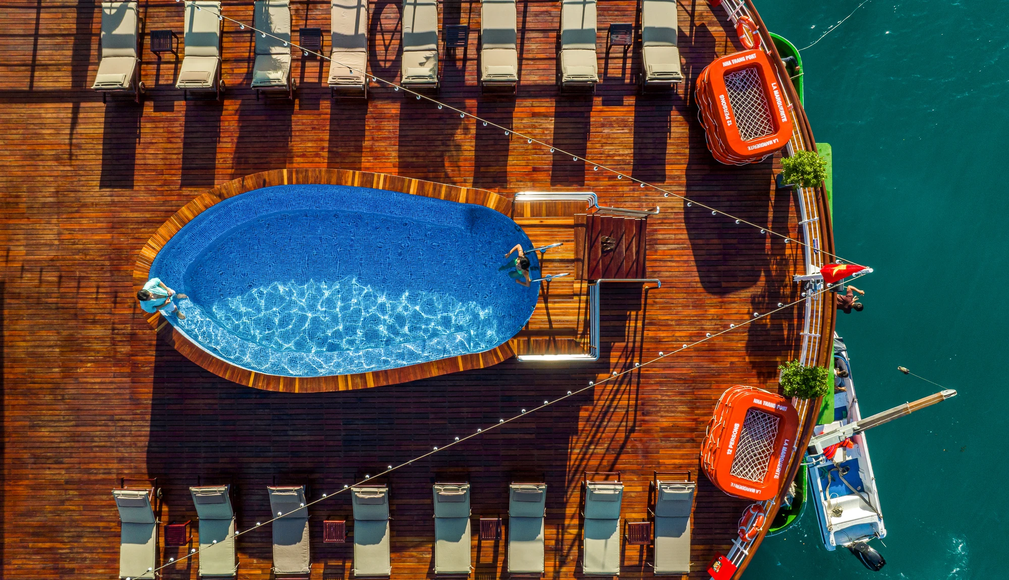 Luftaufnahme eines Poolbereichs mit einem länglichen blauen Pool, umgeben von Holzdecks und Liegen. Zwei orangefarbene Rettungsboote sind sichtbar, sowie ein Boot am Rand.