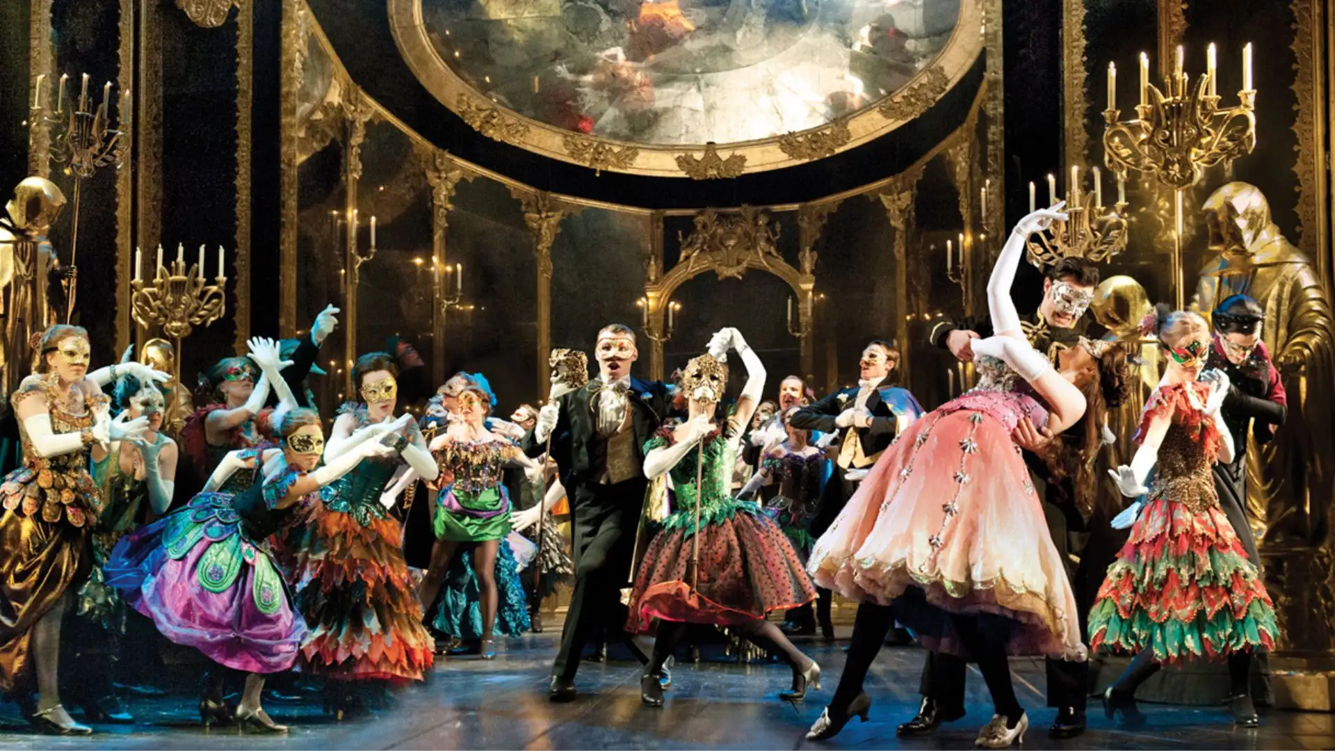 Tanzende Ensemblemitglieder in farbenfrohen Kostümen und Masken auf einer prunkvollen Bühne mit Kronleuchtern und großem Spiegel im Hintergrund in einer Szene des Musicals Phantom der Oper in Köln