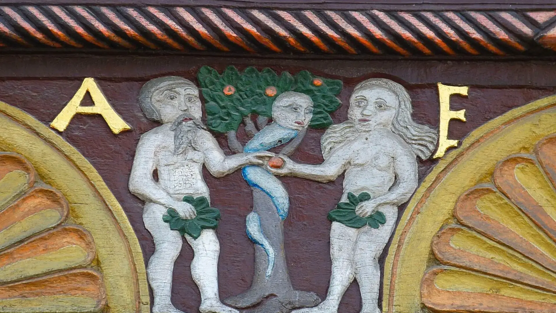 Relief am Adam-und-Eva-Haus in Paderborn zeigt Adam und Eva mit Feigenblättern, Schlange um Baumstamm und goldene Buchstaben 'A' und 'E'.