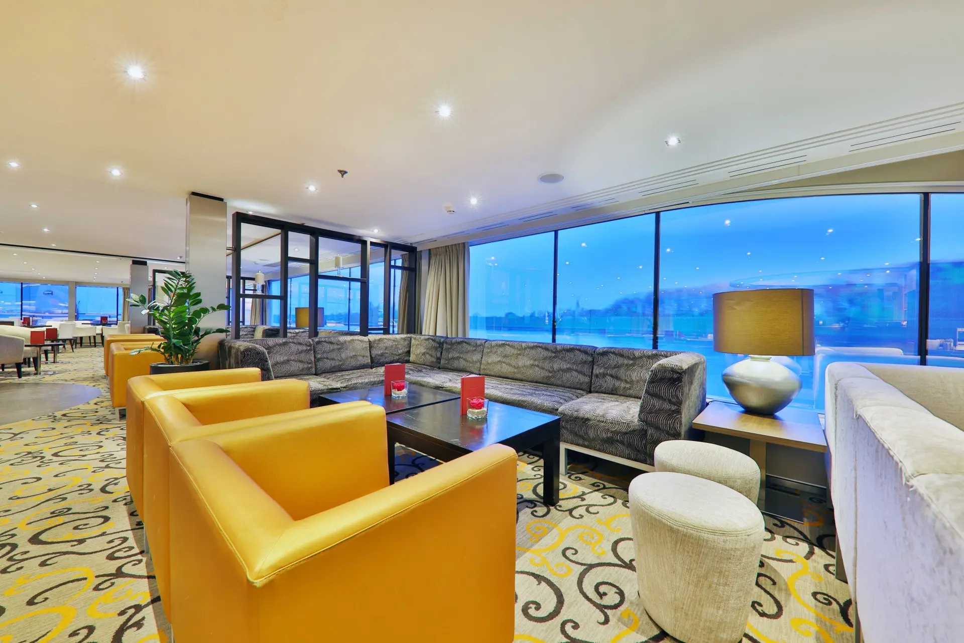 Lounge mit gelben Sesseln, grauem Ecksofa, schwarzen Tischen und großen Fenstern mit Blick auf die Stadt bei Dämmerung