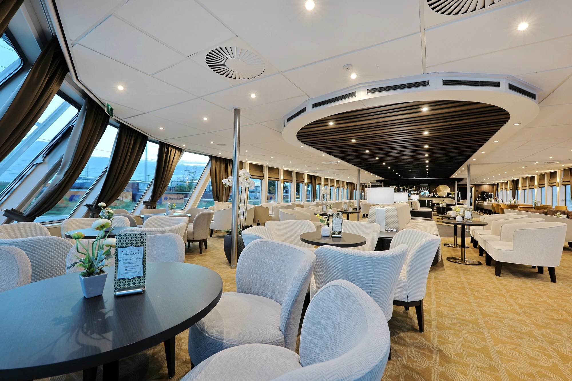 Panoramalounge mit großen Fenstern, runden Tischen und gepolsterten Sesseln in hellem Teppichboden