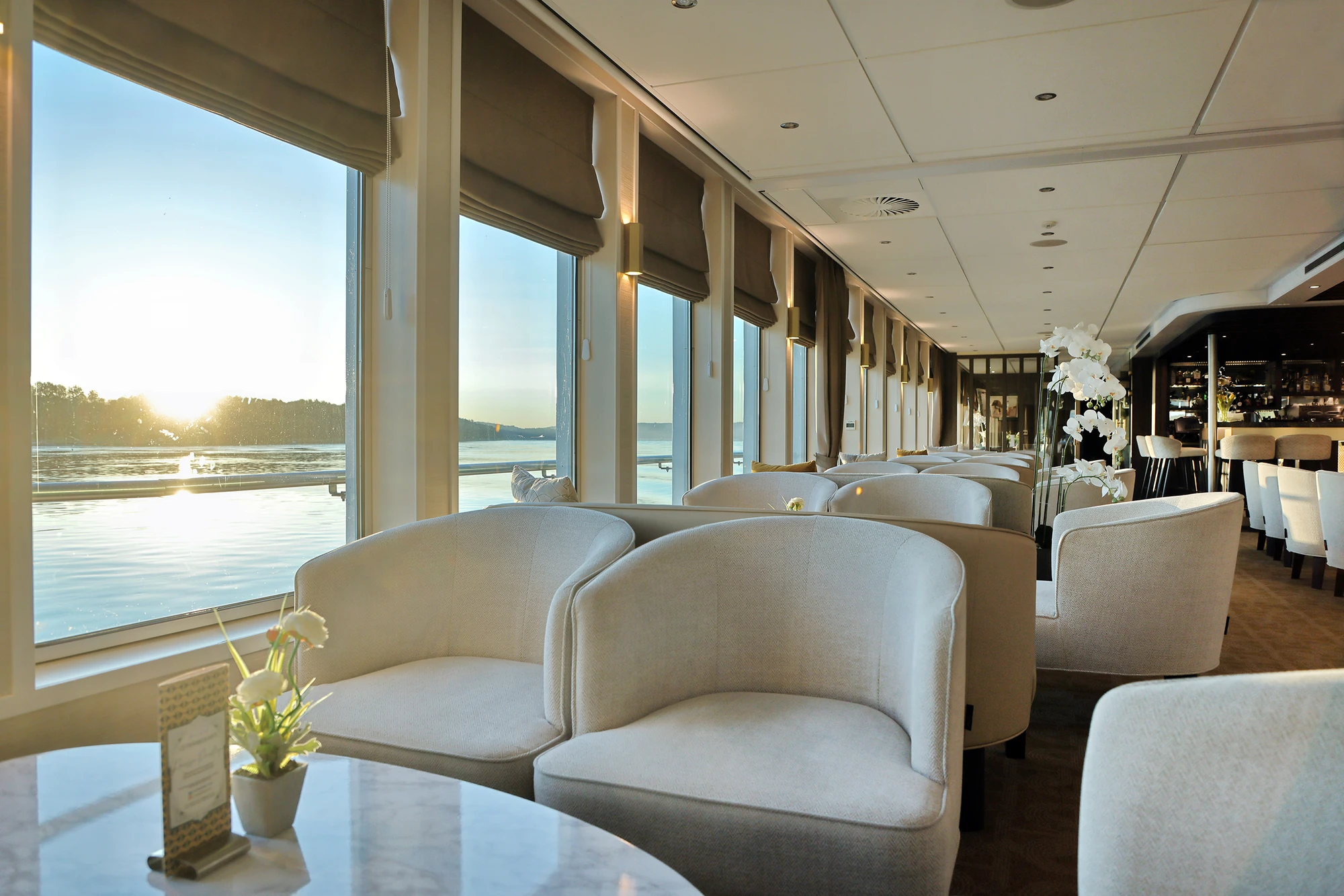 Lounge mit gepolsterten Sesseln und Tischen vor großen Fenstern mit Blick auf einen Fluss bei Sonnenaufgang