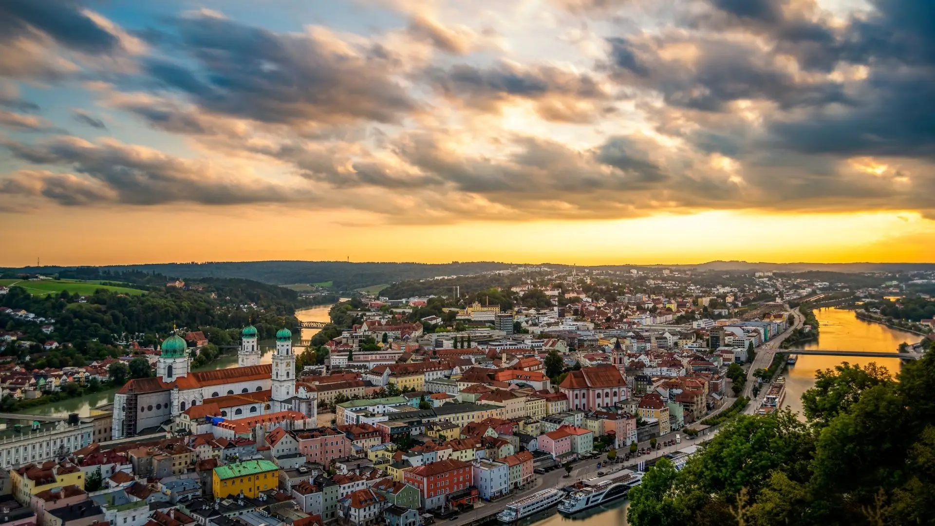 Panoramaansicht von Passau bei Sonnenuntergang, mit der Donau und der Altstadt im Vordergrund, geprägt von bunten Gebäuden und Wolken am Himmel.